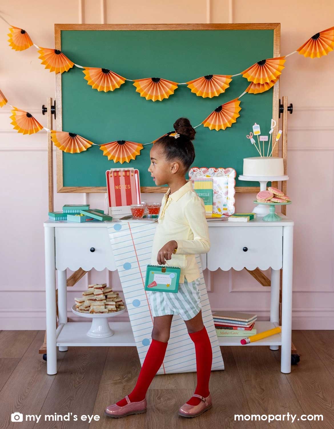 A girl carrying a lunchbox shaped treat box walking in front of a school themed decorated party table with a cake, surrounded by various school themed party supplies and decorations from Momo Party including party plates, napkins, table runner in front of a chalkboard above which is decorated with pencil party fans garland hung above, making this perfect inspiration for kid's first day of school celebration or back to school party.