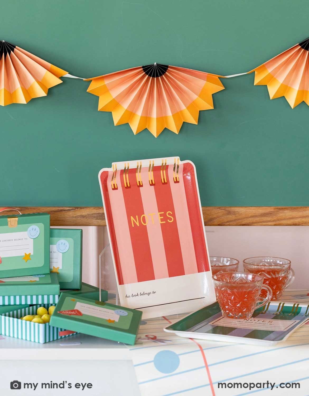 A close up shot of a school themed decorated party table with various school themed party supplies and decorations from Momo Party including red notebook shaped party plate, lunch box shaped treat boxes, and notebook table runner in front of a chalkboard which is decorated with a pencil party fans garland hung above, making this perfect inspiration for kid's first day of school celebration or back to school party.