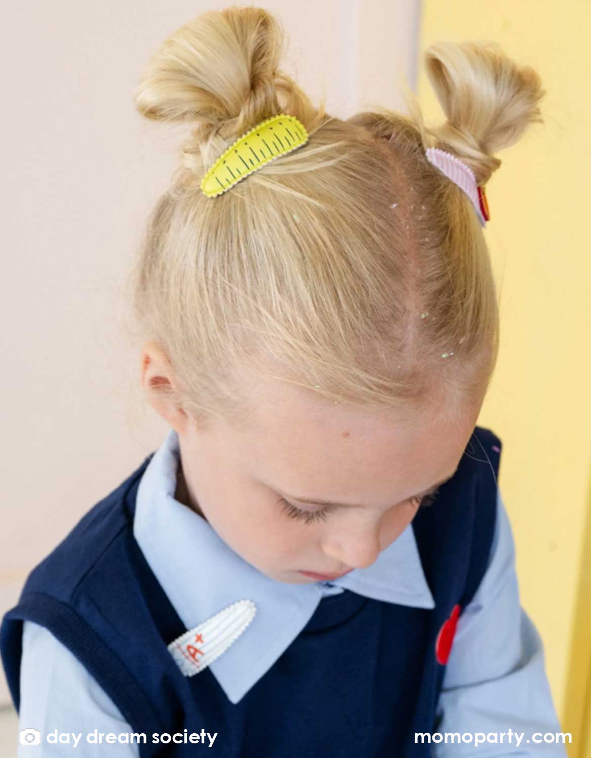 A girl in her uniform with two-bun hair styled with Momo Party's Smarty Pants ABC Hair Clip Set by Daydream Society. Two hair clips, one with a ruler design, another  with an apple design on a red and white stripe background. These hair clips are perfect for back to school picture day, first day of school, and every school day in between.