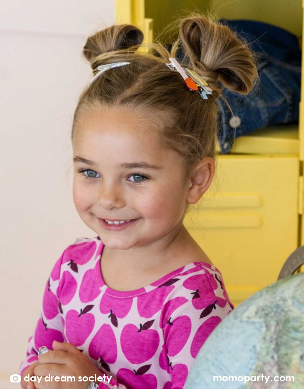 A girl with hair styled in two hair buns adorned with Momo Party's Smarty Pants ABC Hair Bar Clip Set by Daydream Society. Three hair clips, one with alphabet letters A, B, and C, one with pencil design, and one with an apple design. In the back is a classroom set up with yellow lockers. These hair clips are perfect for back to school picture day, first day of school, and every school day in between.