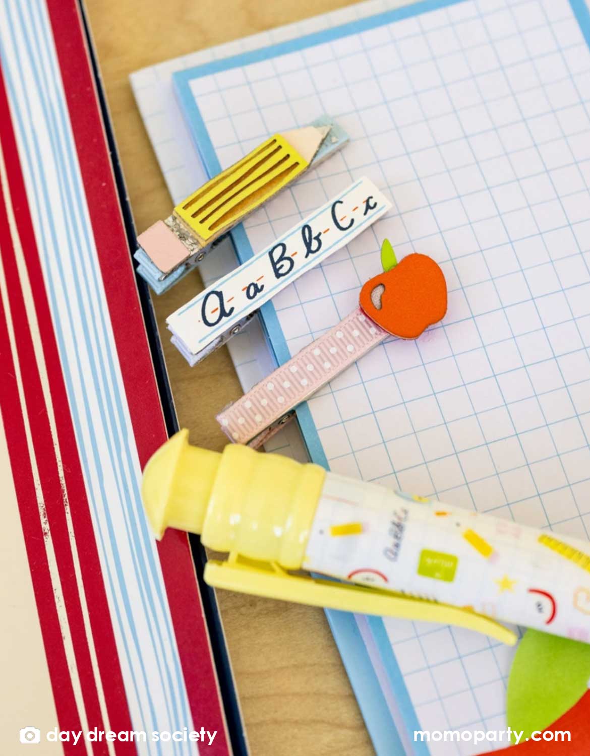 An overhead shot of Momo Party's Smarty Pants ABC Hair Bar Clip Set by Daydream Society. Three hair clips, one with alphabet letters A, B, and C, one with pencil design, and one with an apple design. These hair clips are surrounded by school supplies including grid notebooks, pens, and books.