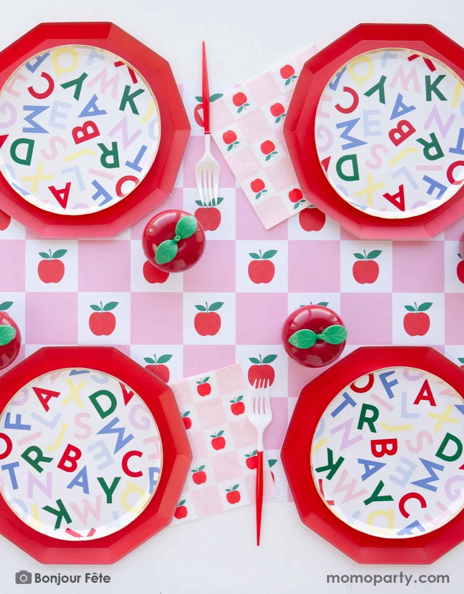 A festive back to school apple themed party table setting featuring various apple and school themed party tableware from Momo Party including large red dinner plates, alphabet side plates, red and white utensils, apple and pink checkered table runner and matching large napkins. With some cute apple shaped centerpiece in the middle, this makes a perfect tablescape for kid's back to school party or first day of school celebration.