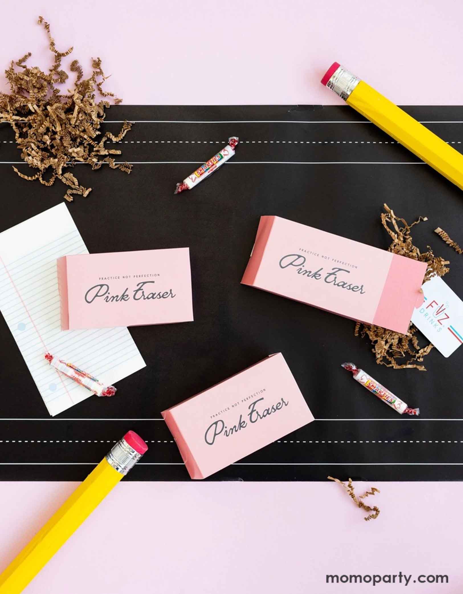 An overhead shot of a back to school party table featuring various school themed party supplies from Momo Party including pink eraser shaped treat boxes, notebook shaped napkins, and chalk board paper table runner. Around the party supplies are some smarty candies, a couple of jumbo pencils as props -  this table settings is a perfect inspiration for a simple yet adorable idea for kid's goodie bags at a school themed event or celebration.