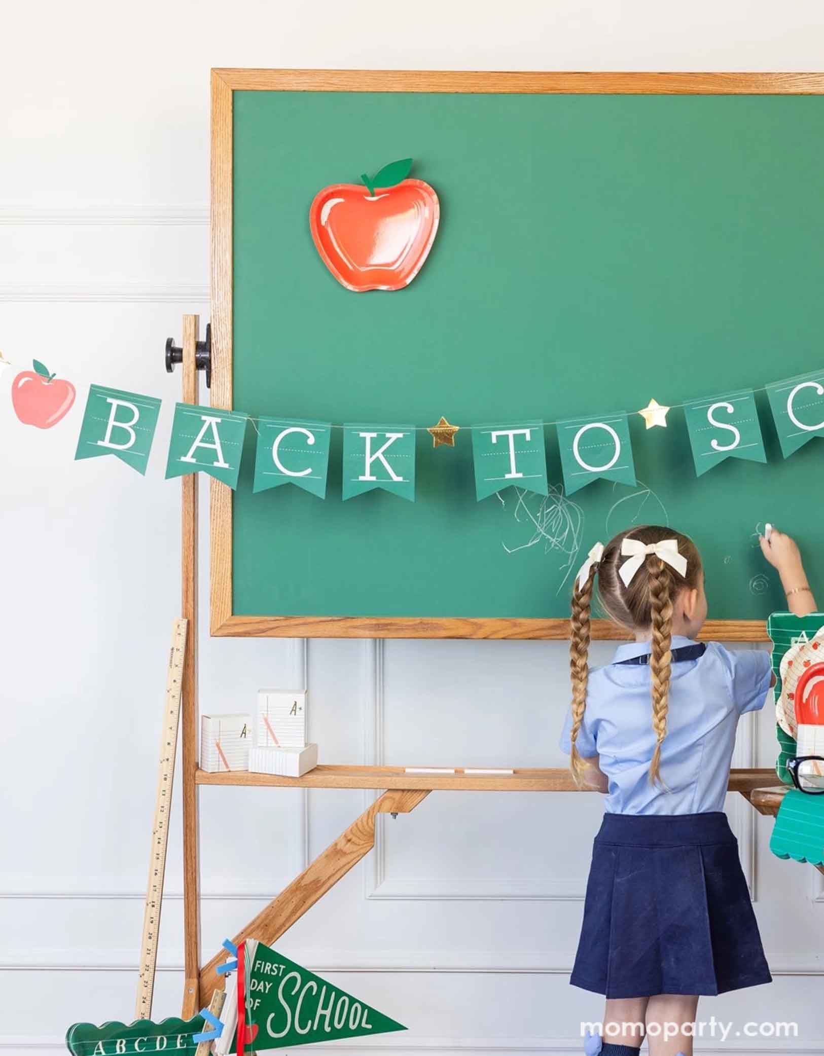 A school-aged girl in her blue uniform writing something on a chalkboard in a classroom set up decorated with a bunch of school themed party supplies from Momo Party including 8.5" apple shaped plates, 10" apple pattern plates, alphabet dinner plates and matching napkins. The chalkboard is adorned with the back to school party banner hung across, with an apple shaped plate on it - an adorable and cute set up to celebrate kid's back to school season!