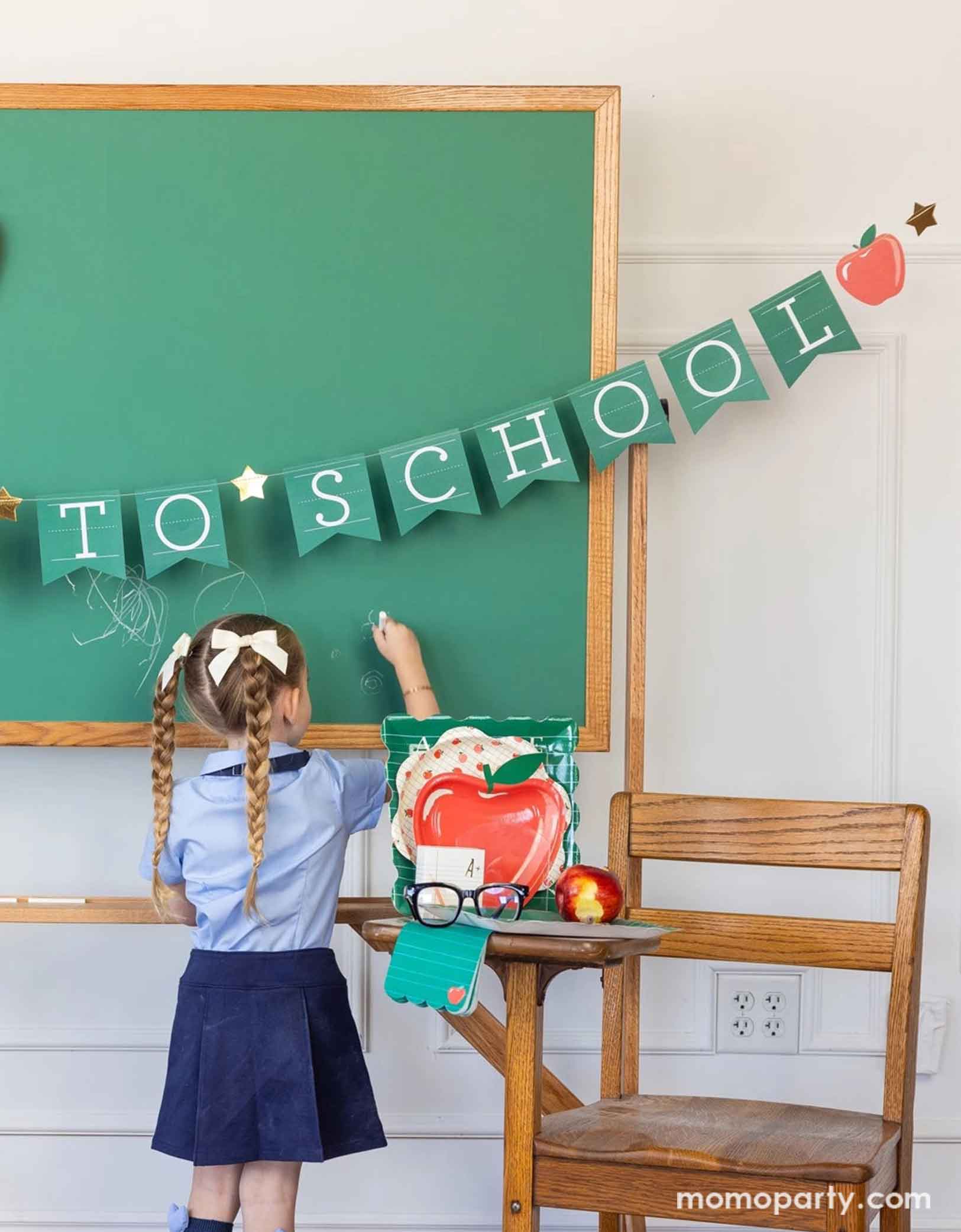 A school-aged girl in her blue uniform writing something on a chalkboard in a classroom set up decorated with a bunch of school themed party supplies from Momo Party including 8.5" apple shaped plates, 10" apple pattern plates, alphabet dinner plates and matching napkins. The chalkboard is adorned with the back to school party banner hung across, with an apple shaped plate on it - an adorable and cute set up to celebrate kid's back to school season!
