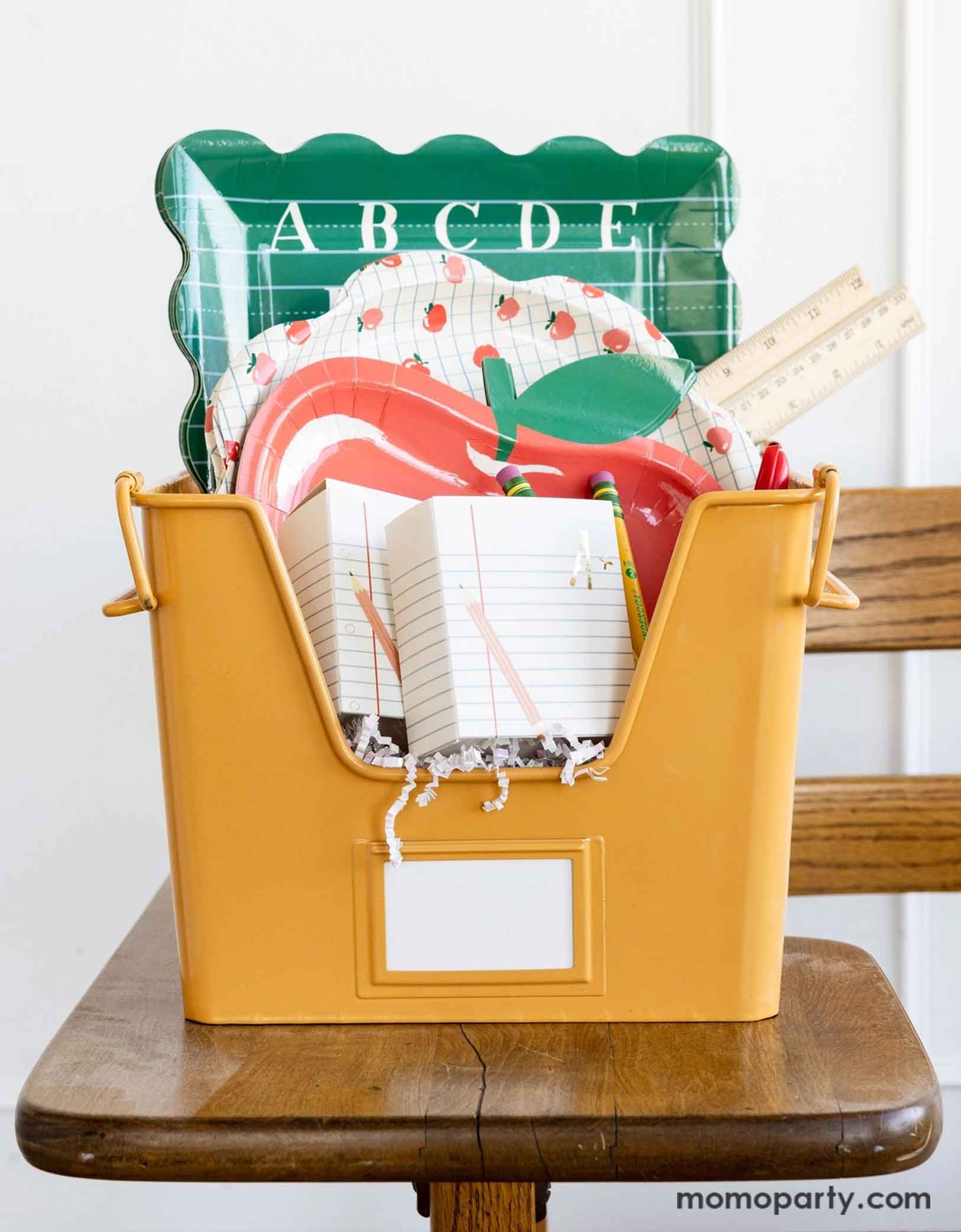 A vintage inspired yellow file bin filled with various school themed party supplies from Momo Party's including two ruled paper treat boxes, an apple shaped paper plate, an apple patterned scallop-edged plate and an ABC alphabet green dinner plate. The bin is placed on a classic classroom chair - a perfect inspiration for celebrating kid's back to school season or first day of school occasion!
