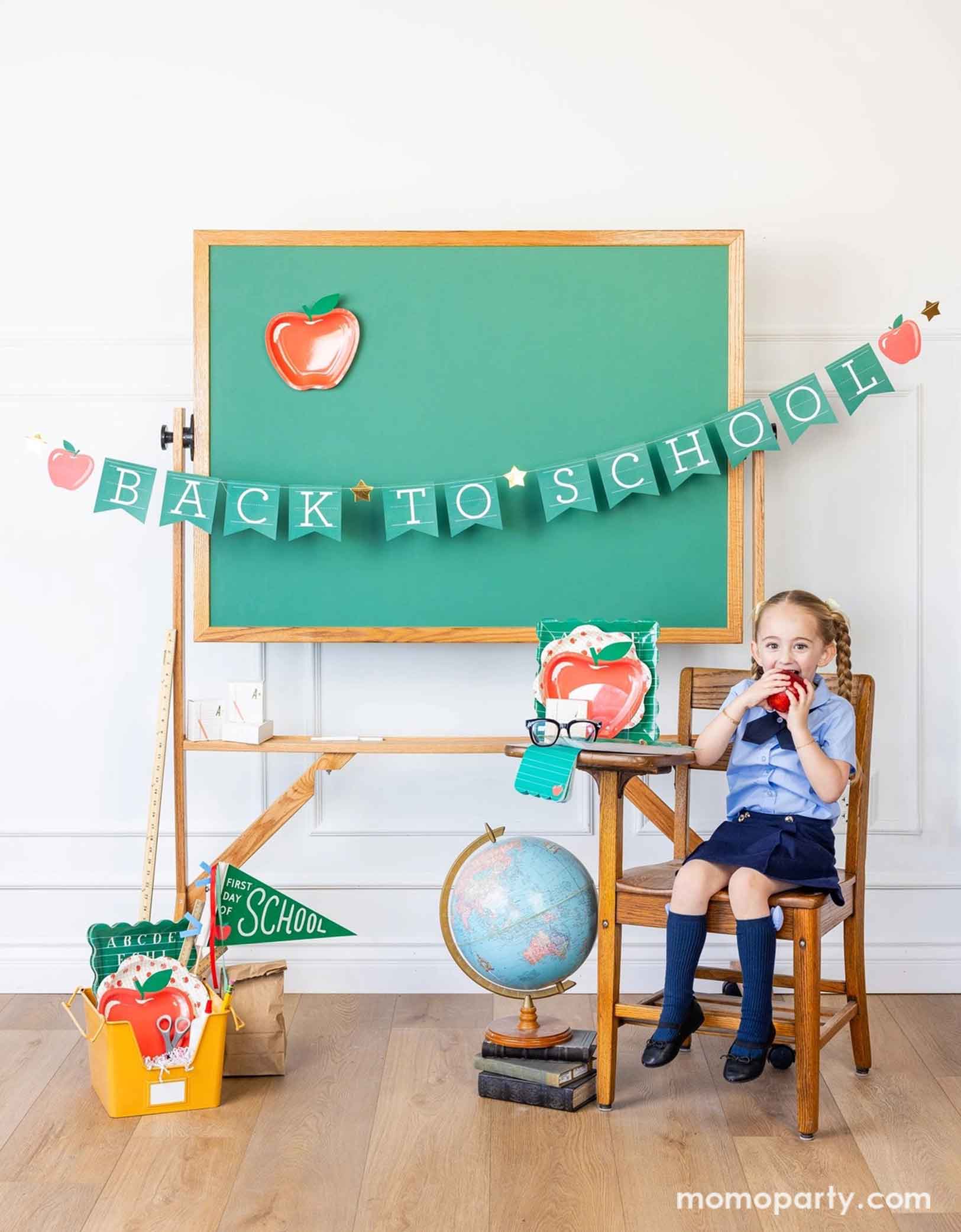 A school-aged girl in her blue uniform eating an apple on a school chair in a classroom setting decorated with a bunch of school themed party supplies from Momo Party including 8.5" apple shaped plates, 10" apple pattern plates, alphabet dinner plates and matching napkins. Behind her is a classroom chalkboard with back to school party banner hung across with an apple shaped plate on it - an adorable and cute set up to celebrate kid's back to school season!