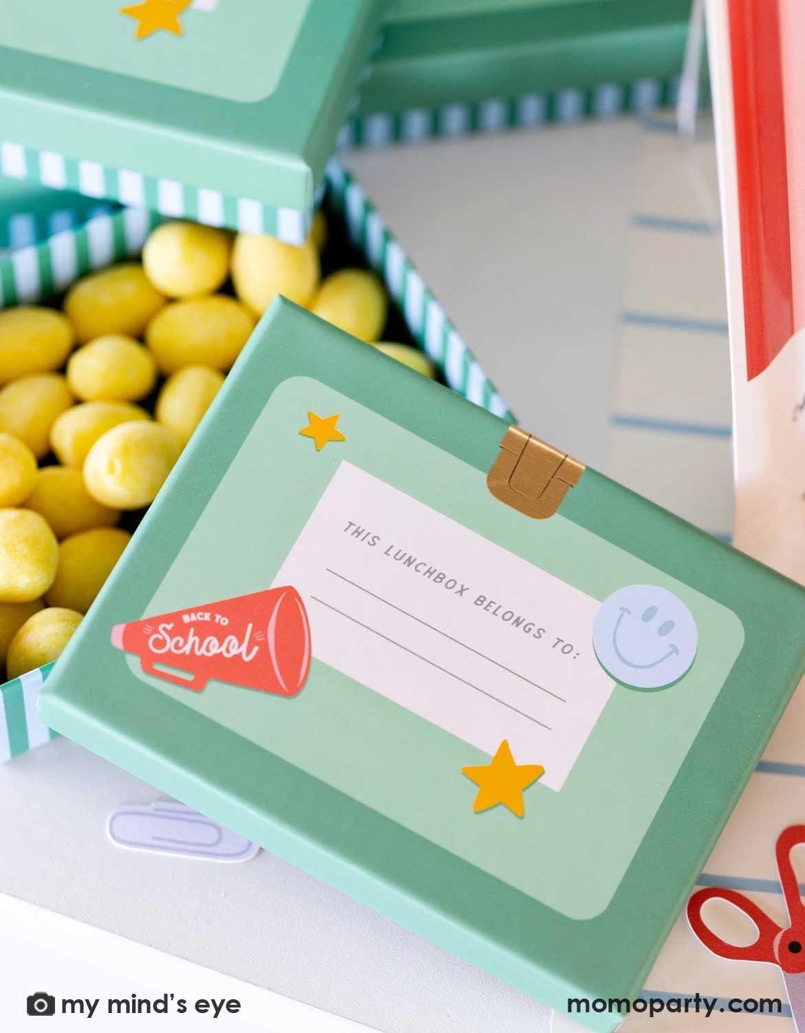 A close up shot of Momo Party's lunch box shaped treat box filled with yellow candies on a school themed party table featuring various of school themed party supplies from Momo Party.