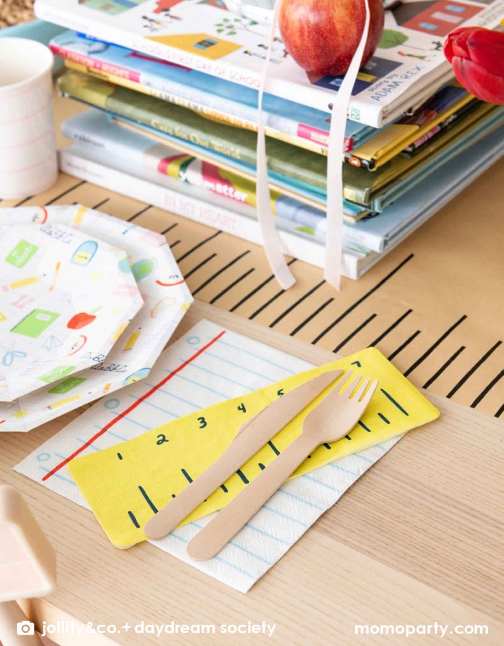 A close up shot of an adorable kid's school themed party table featuring school supplies themed party tablewares from Momo Party including the Smarty Pants ruler shaped napkins and notebook shaped napkins along with large and small school icons patterned plates, all on a ruler inspired table runner. In the middle of the table there is a pile of books and some apples as centerpiece, making this a great decoration inspiration for kid's  first day of school celebration or a back to school party.
