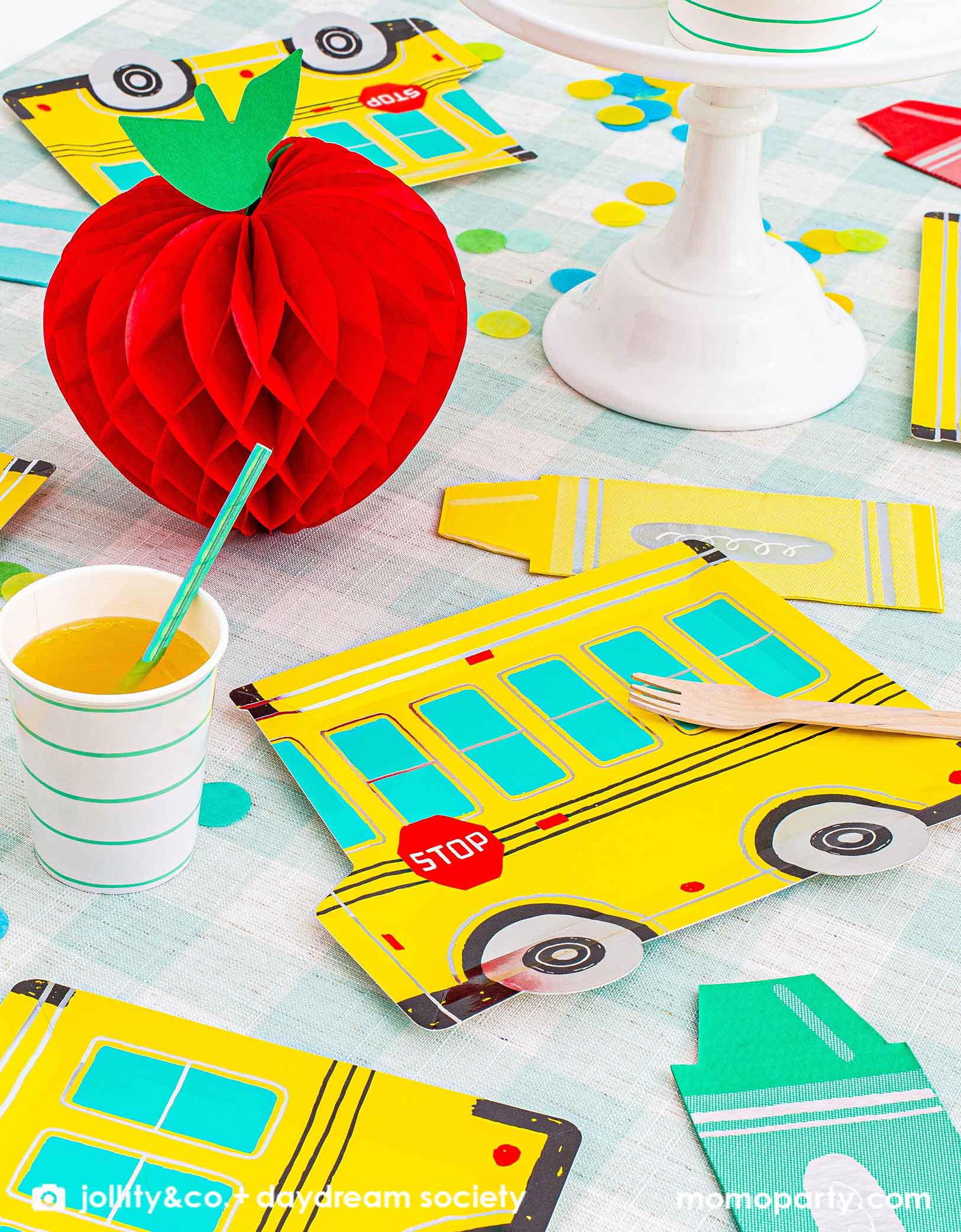 A festive back to school party table featuring Momo Party's colorful school themed tableware including Daydream Society's school bus shaped plates, crayon shaped napkins with aqua striped party cups, all on a mint gingham checkered tablecloth. With a festive apple shaped honeycomb decoration on the party table and some bright colored confetti spread out, this gives a great idea and inspiration for a kid's back to school party or a first day of school celebration.