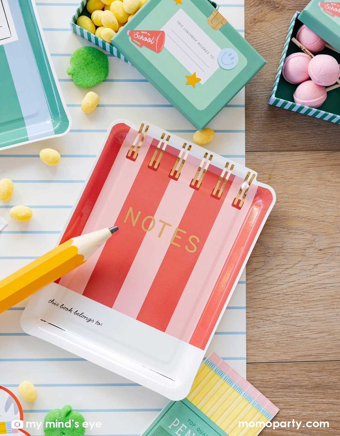 An overhead shot of a school themed party table featuring Momo Party's red striped notebook shaped plate, pencil set napkins and small lunch box shaped treat boxes filled with candies, with green apple shaped gummies and cupcakes spread across the notebook blue line paper table runner, making this a perfect back to school party or first day of school celebration tablescape inspiration.