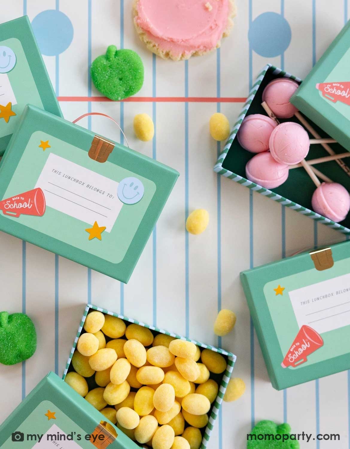 An overhead shot of a school themed party table featuring Momo Party's lunch box shaped treat boxes filled with colorful candies and lollipops, with green apple shaped gummies and cupcakes spread across the notebook blue line paper table runner, making this a perfect tablescape inspiration for kid's school themed party or first day of school celebration.