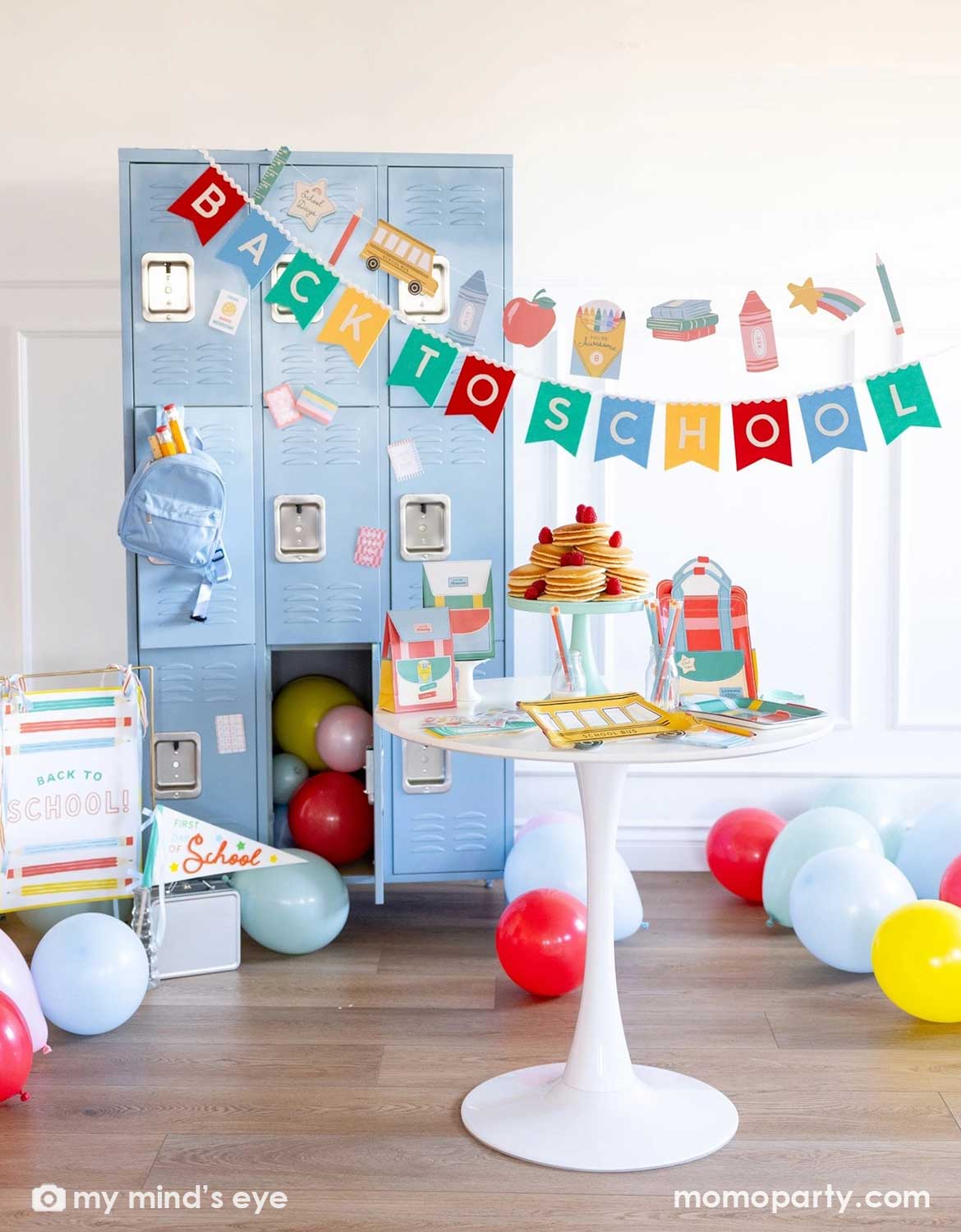 A back to school party set up featuring various school themed party supplies and decors from Momo Party including school themed plates, treat boxes, party pennant, back to school felt garland along with school icons party banner hung above the light blue locker selves. A party table with pancakes on a cake stand next to some colorful backpack shaped treat bags. Along with colorful balloons spread across the room, this is a perfect inspiration for decorating ideas for kids first day of school celebration.