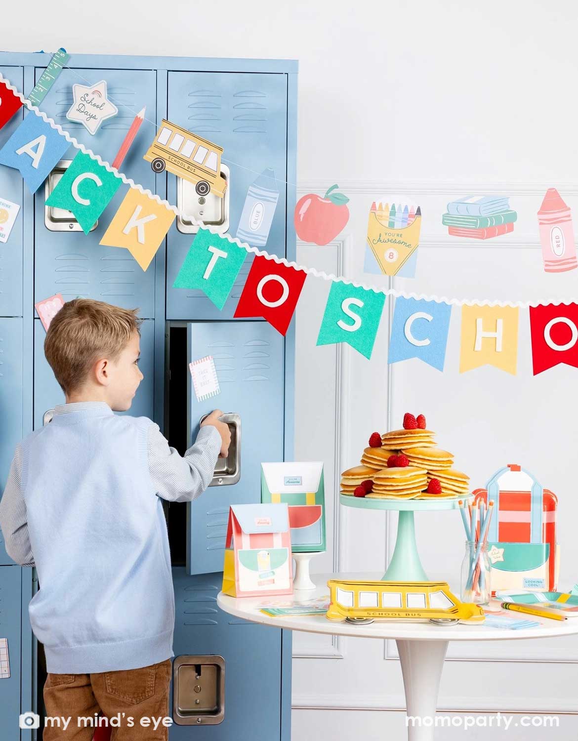 A boy opening a blue locker in a back to school classroom set up featuring various school themed party supplies and decors from Momo Party including school themed plates, treat boxes, party pennant, back to school felt garland along with school icons party banner hung above the light blue locker selves. A party table with pancakes on a cake stand next to some colorful backpack shaped treat bags.
