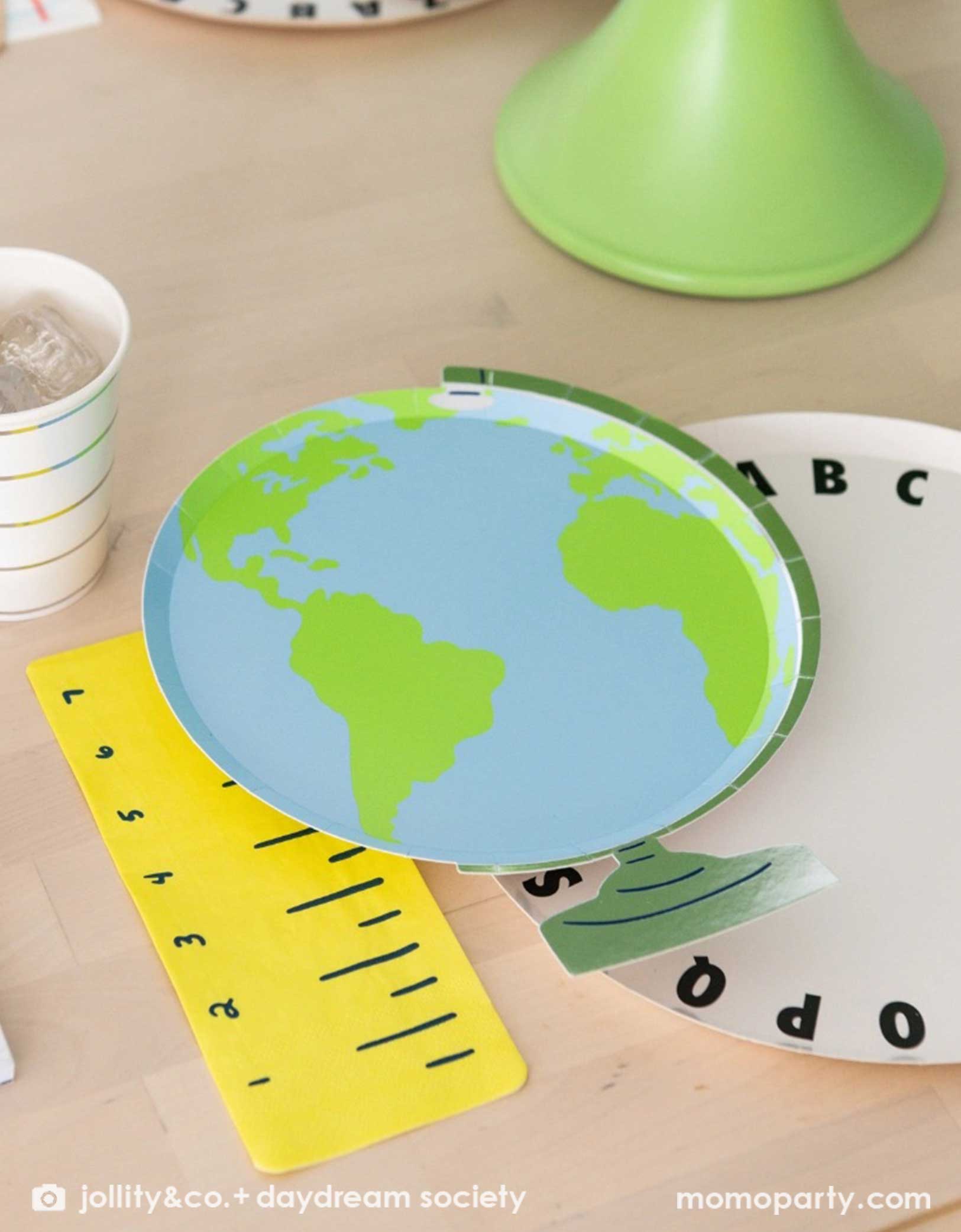 A close up shot of an adorable kid's school themed party featuring school supplies themed party tablewares from Momo Party including the Smarty Pants Globe shaped Large Plate, alphabet round dinner plate, ruler shaped napkin and striped party cup - making this a cute and festive tablescape for kid's school themed celebration, be it the last day of school party, the first day of school celebration or a back to school party.