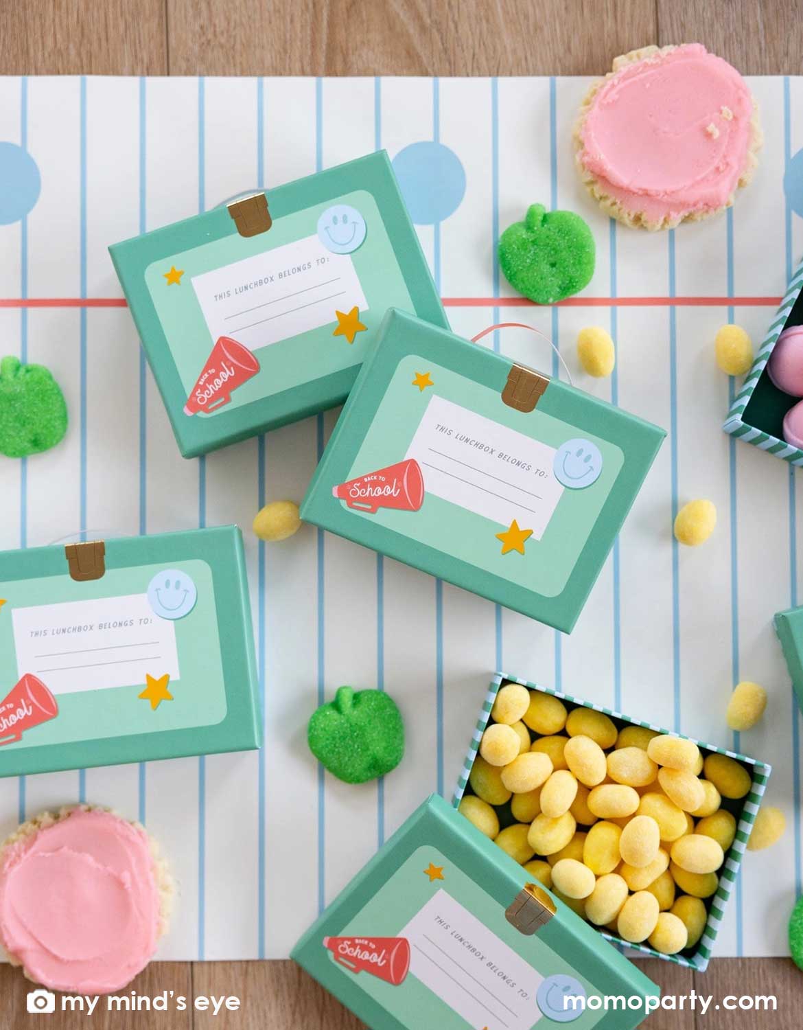 An overhead shot of a school themed party table featuring Momo Party's lunch box shaped treat boxes filled with colorful candies and lollipops, with green apple shaped gummies and cupcakes spread across the notebook blue line paper table runner, making this a perfect tablescape inspiration for kid's school themed party or first day of school celebration.