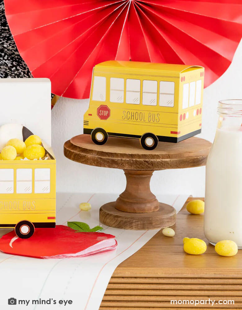 A festive back to school party table features Momo Party's school themed party supplies including school bus shaped treat boxes, apple shaped napkins, writing paper table runner by My Mind's Eye. On the table are some red jelly beans, yellow candies and milk bottles in coordinating colors. In the back there's the back to school party fan hung on the wall, making this a perfect inspiration for kid's back to school party or a first day of school celebration breakfast.