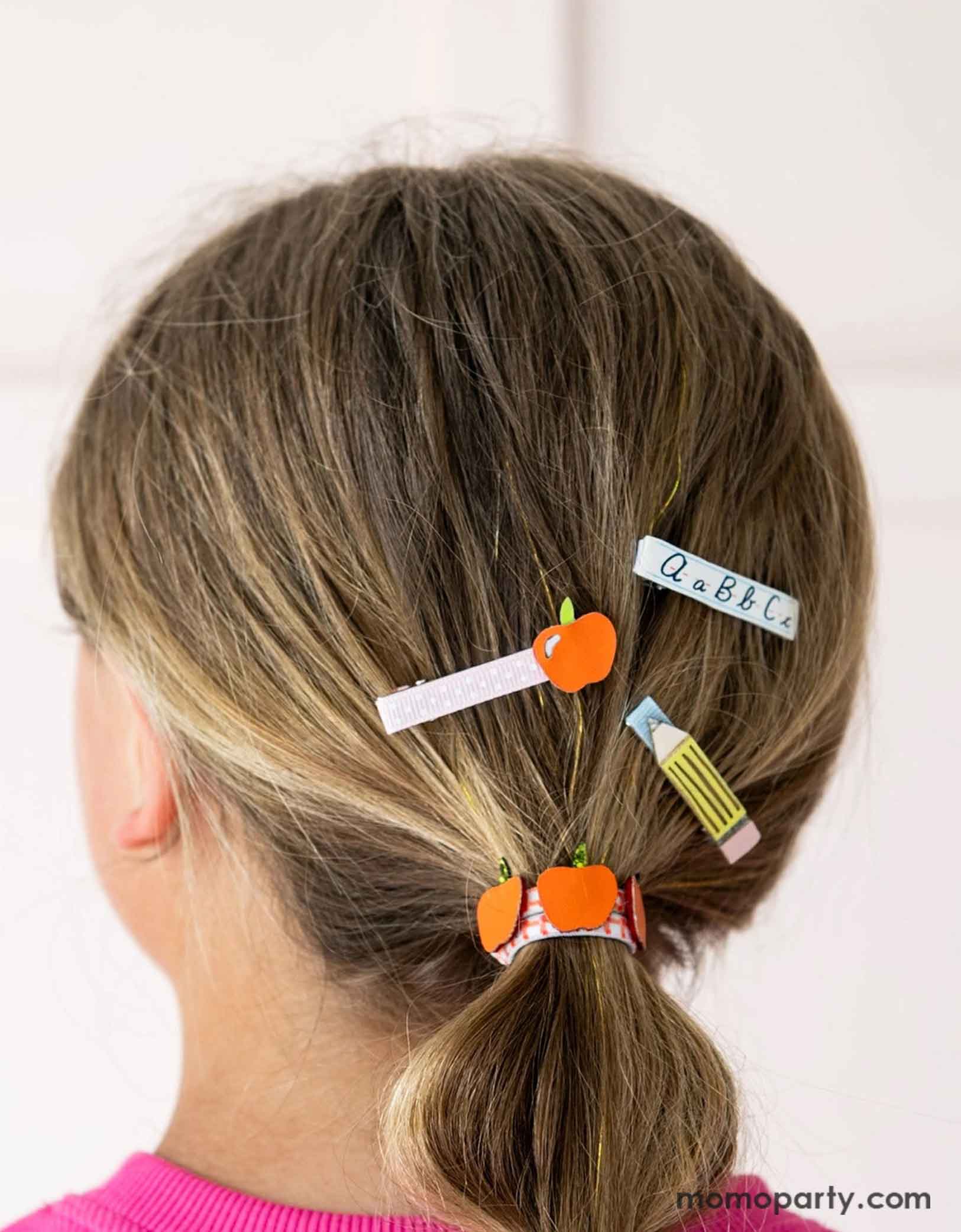 A girl with hair styled in a ponytail using Momo Party's Smarty Pants ABC Hair Bar Clip Set by Daydream Society. Three hair clips, one with alphabet letters A, B, and C, one with pencil design, and one with an apple design. These hair clips are perfect for back to school picture day, first day of school, and every school day in between.