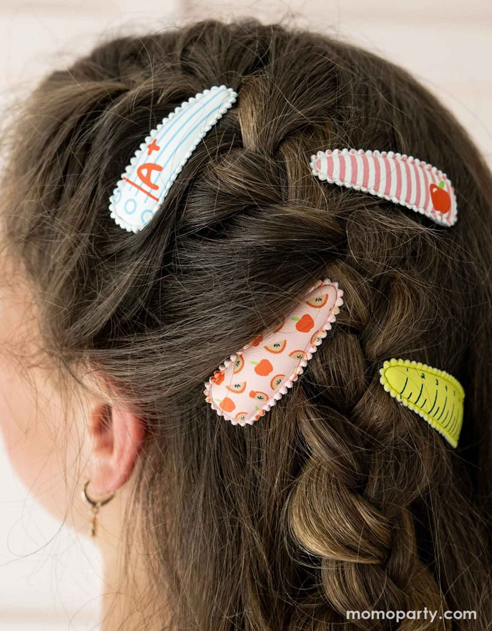 A girl with hair styled in braids using Momo Party's Smarty Pants ABC Hair Clip Set by Daydream Society. Four hair clips, one with a notebook design with A+ on it, one with an apple design on a red and white stripe background, one with apple and rainbow patterned design, and one with a ruler design. These hair clips are perfect for back to school picture day, first day of school, and every school day in between.