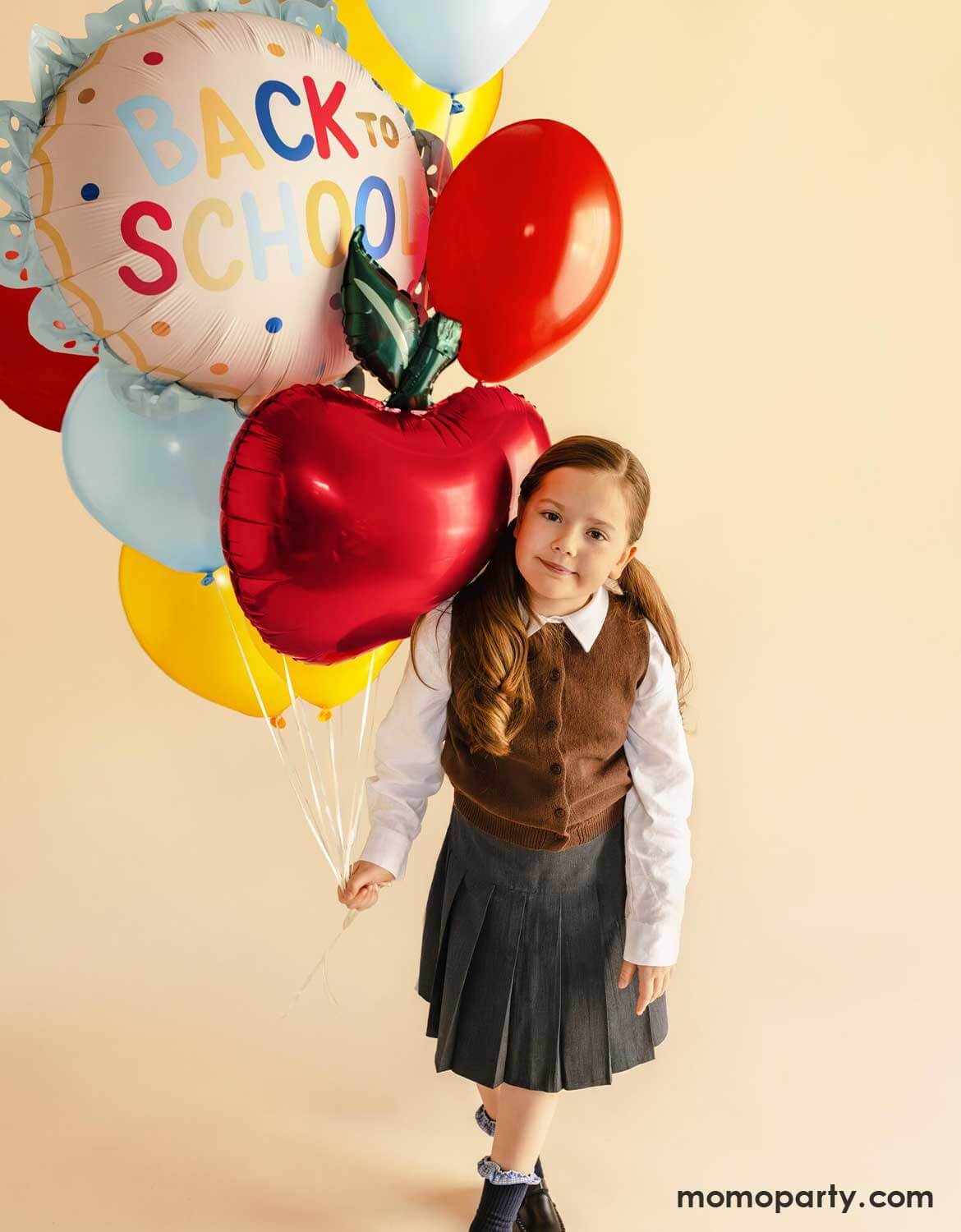 A young girl in her uniform holding a balloon bouquet which features Momo Party's 20" scalloped "Back to School" foil balloon, an apple shaped foil balloon and a bunch of latex balloons in festive colors against a beige background.