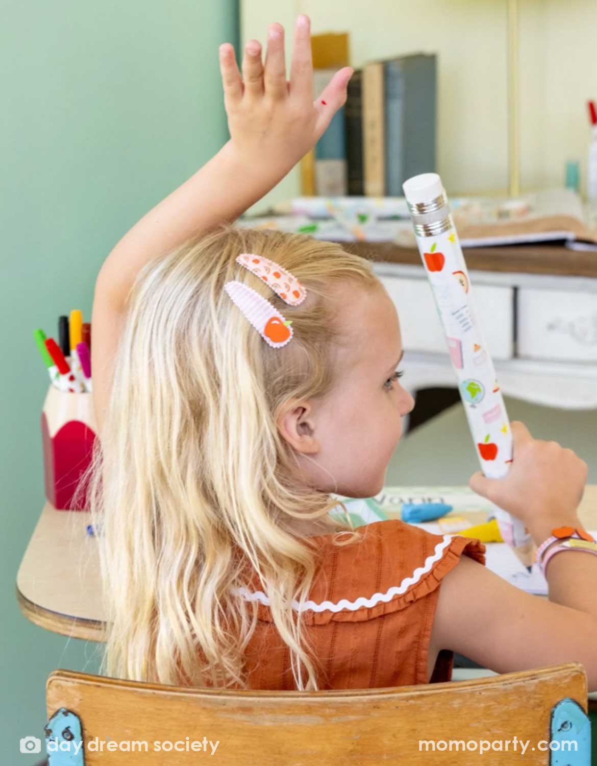 A girl in a classroom setting sitting on a school desk raising her hand. Her hair is styled with Momo Party's school themed hair clips in apple design. She's also holding a giant pencil with school themed designs on it. These hair clips are perfect for back to school picture day, first day of school, and every school day in between.