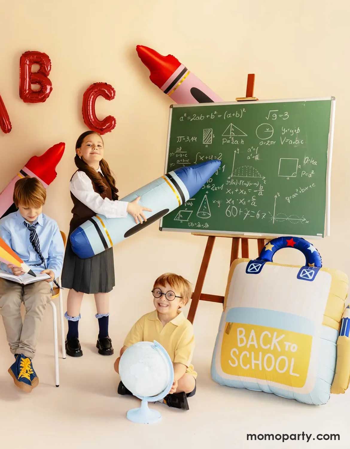 Children in their uniform in school-themed first day of school setting with a chalkboard, crayons shaped balloons in blue and red, and 'ABC' foil balloons on the wall, along with Momo Party's backpack shaped foil balloon with "Back to School" text on it.