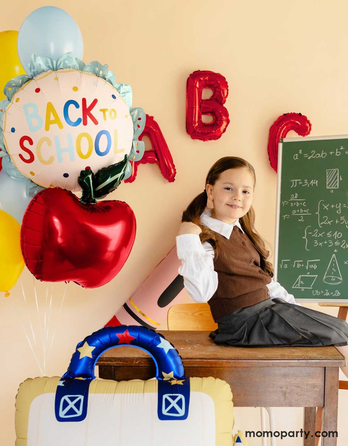 A young girl in her uniform sitting in a classroom setting with Momo Party's 20" scalloped "Back to School" foil balloon, an apple shaped foil balloon along with red alphabet balloons on the wall next to a chalkboard - a perfect setup for kids back to school party, classroom decorations or first day of school celebrations.
