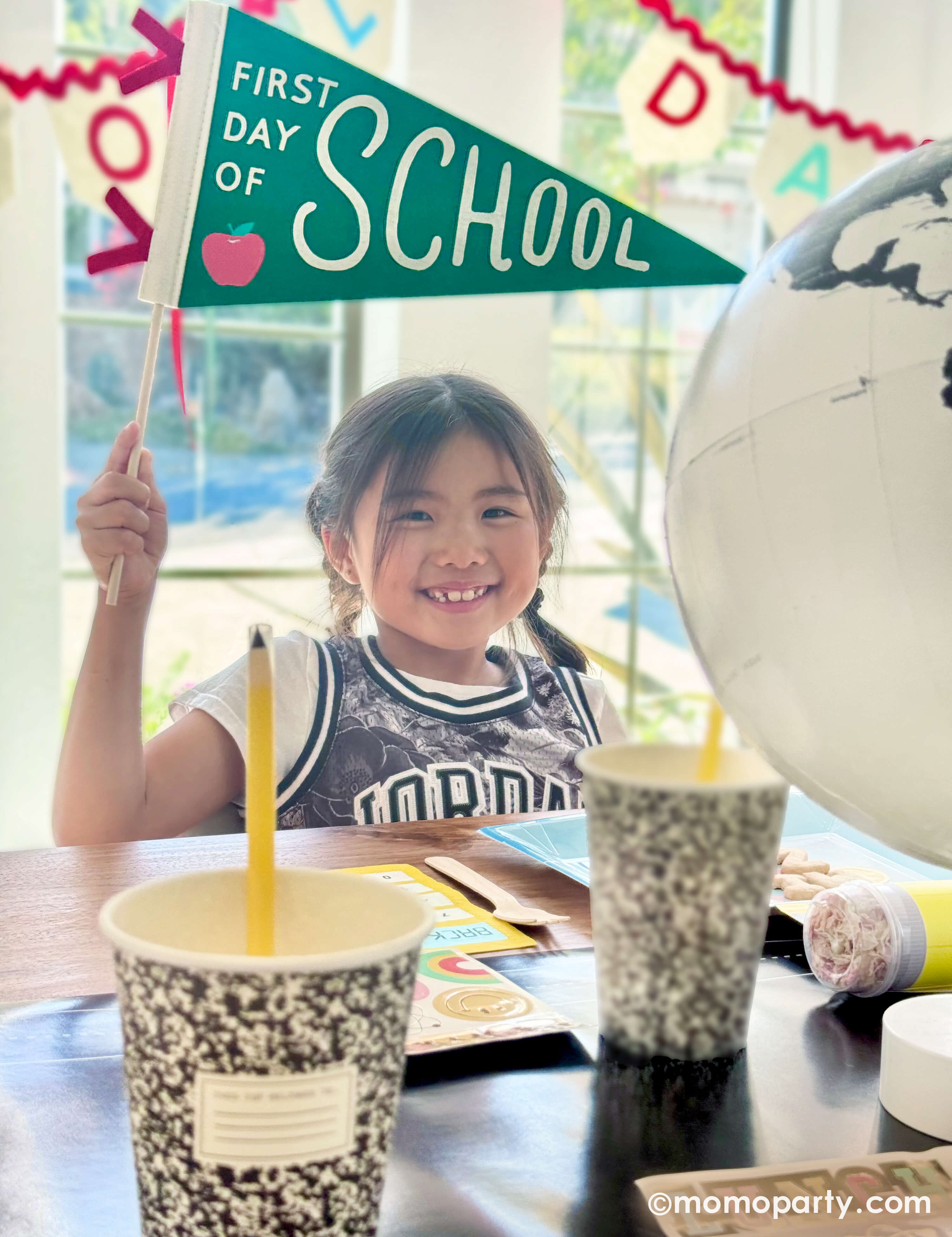 A school aged girl at a back to school celebration holding Momo Party's first day of school apple felt party banner sitting at a table decorated with various of school themed decorations and party tablewares from Momo Party including the chalkboard table runner, notebook party cups, pencil confetti poppers, lunchbox shaped party plates and school day felt party garland. A perfect inspo for your child's first day of school celebration!