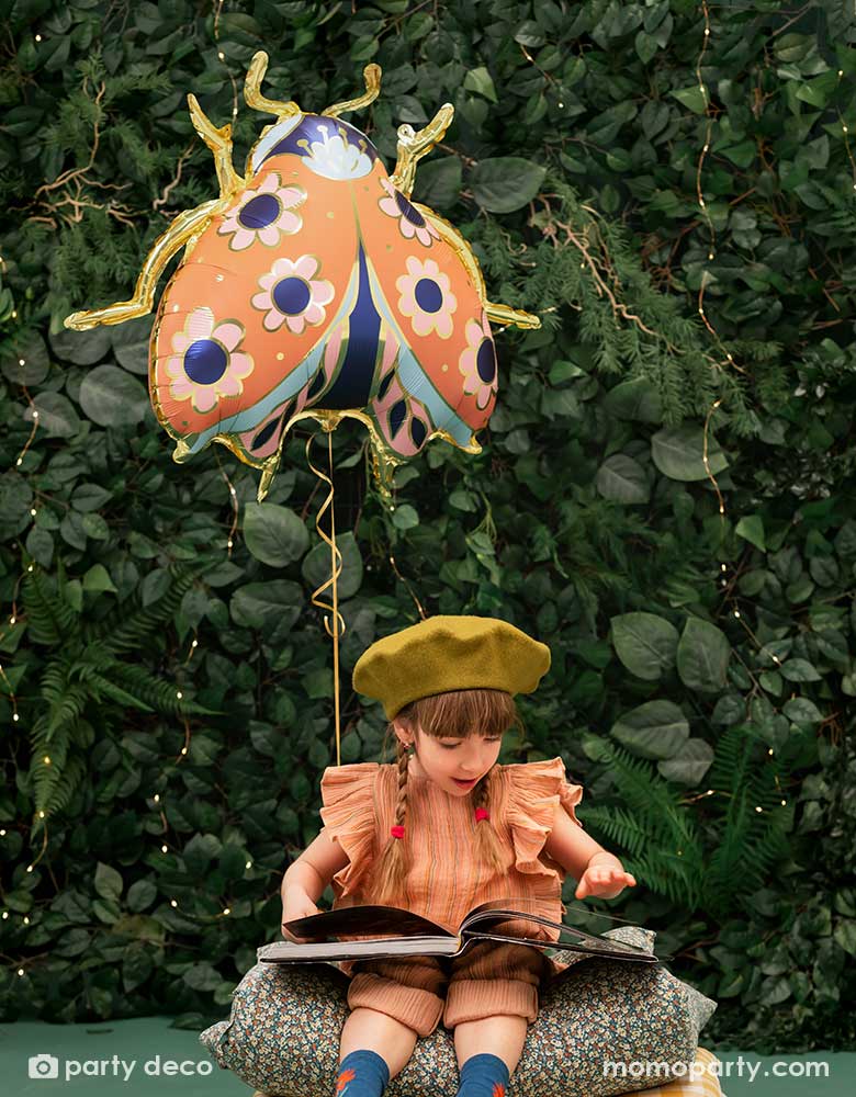 A little girl reading on her book in the backyard with a wall of greenery, next to her is Momo Party's 34 x 30 inches ladybird shaped foil balloon by Party Deco. With coral color with small flower pattern with gold legs on it.