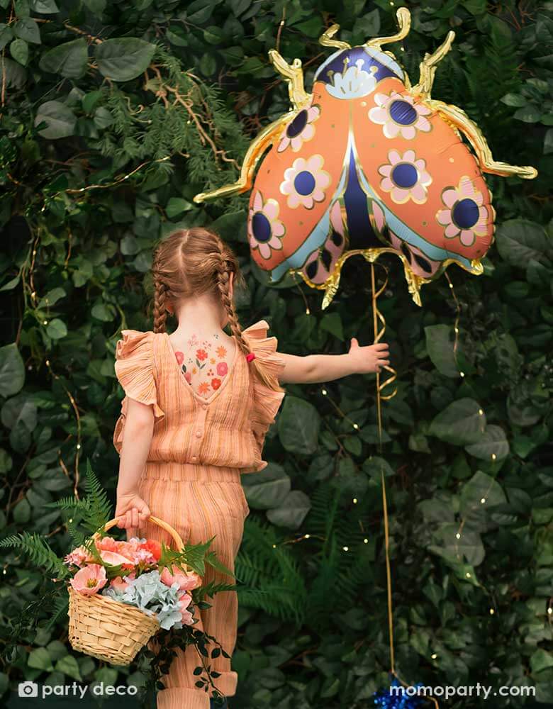 A little girl holding Momo Party's 34 x 30 inches ladybird shaped foil balloon by Party Deco. With coral color with small flower pattern with gold legs on it. She's dressed in her spring outfit holding a basket of spring flowers standing in a backyard with a wall filled of greenery.