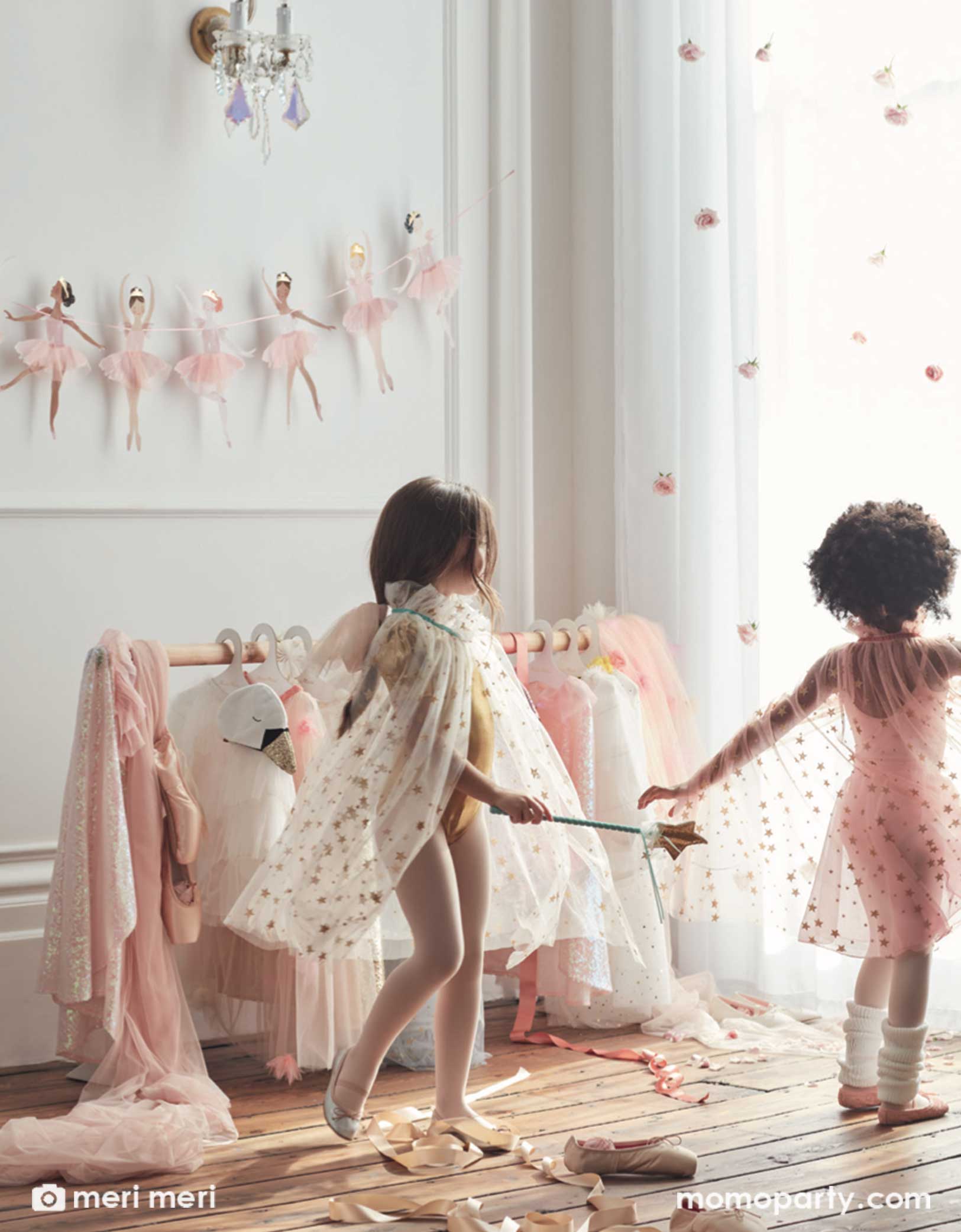 Two girls dressed in tutus in a Ballet themed birthday party. On the wall there's Meri Meri's Ballerina Garland hung on the wall. There are ribbons and a rack of tutus next to the girls making this a dreamy set up  for a Ballerina birthday party, girl birthday party, or a princess party.