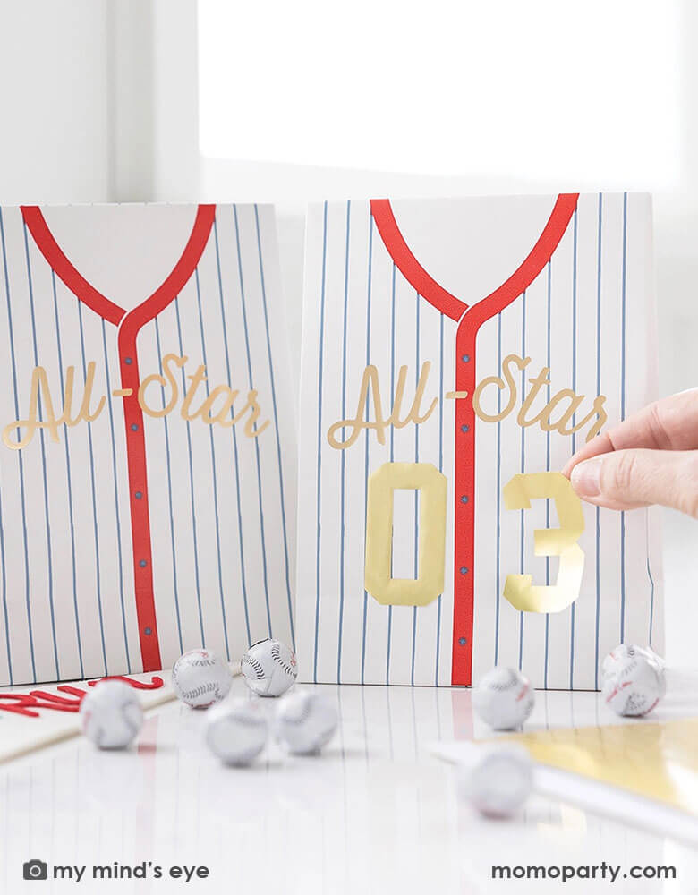 A baseball party table features Momo Party's baseball treat bags by My Mind's Eye. A hand is putting the gold number stickers to the bad to personalize the favor bag. On the table there are some baseball chocolate balls. With these treat bags, they make great party favor for your kid's friends at their baseball themed party!