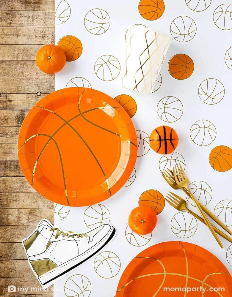 A basketball themed party table features Momo Party's basketball tableware by My Mind's Eye including basketball shaped plates, hightop sneakers shaped napkins, basketball net gold foil party cups and paper basketball table runner, with oranges as snacks on the table - a perfect table setting inspo for kid's basketball themed birthday parties or a fun NBA viewing party!