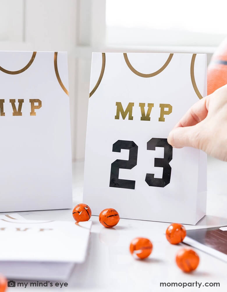 A basketball themed party table features Momo Party's basketball treat bags by My Mind's Eye. Comes in a set of 8 treat bags, these basketball jersey shaped treat bags with MVP on them in gold foil are personalizable with the black number stickers. On the table there are some basketball chocolate balls, makes this a perfect idea for party favors at kid's basketball themed birthday party. 