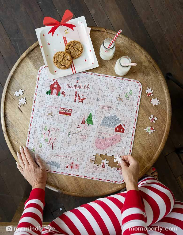 A lady in her red and white striped pajama playing a Christmas themed puzzle next to Momo Party's Believe Santa Present with a bow paper plates with cookies in them, next to it are two bottles with milk with red striped straws, creating a Holiday cozy vibe at home.