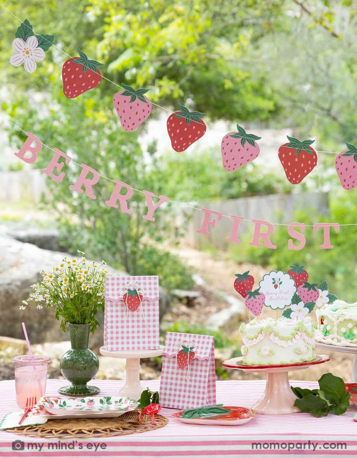 A Berry First strawberry-themed first birthday party setup outdoors, featuring various strawberry themed party supplies from Momo Party including a pink and red strawberry banner garland, a 'BERRY FIRST BIRTHDAY' banner, strawberry cake topper decorations on a vintage strawberry butter cake, and gingham party favor bags with strawberry cutouts, along with strawberry themed tablewares like plates, napkins, cups and flowers on a table with a blurred green garden background.