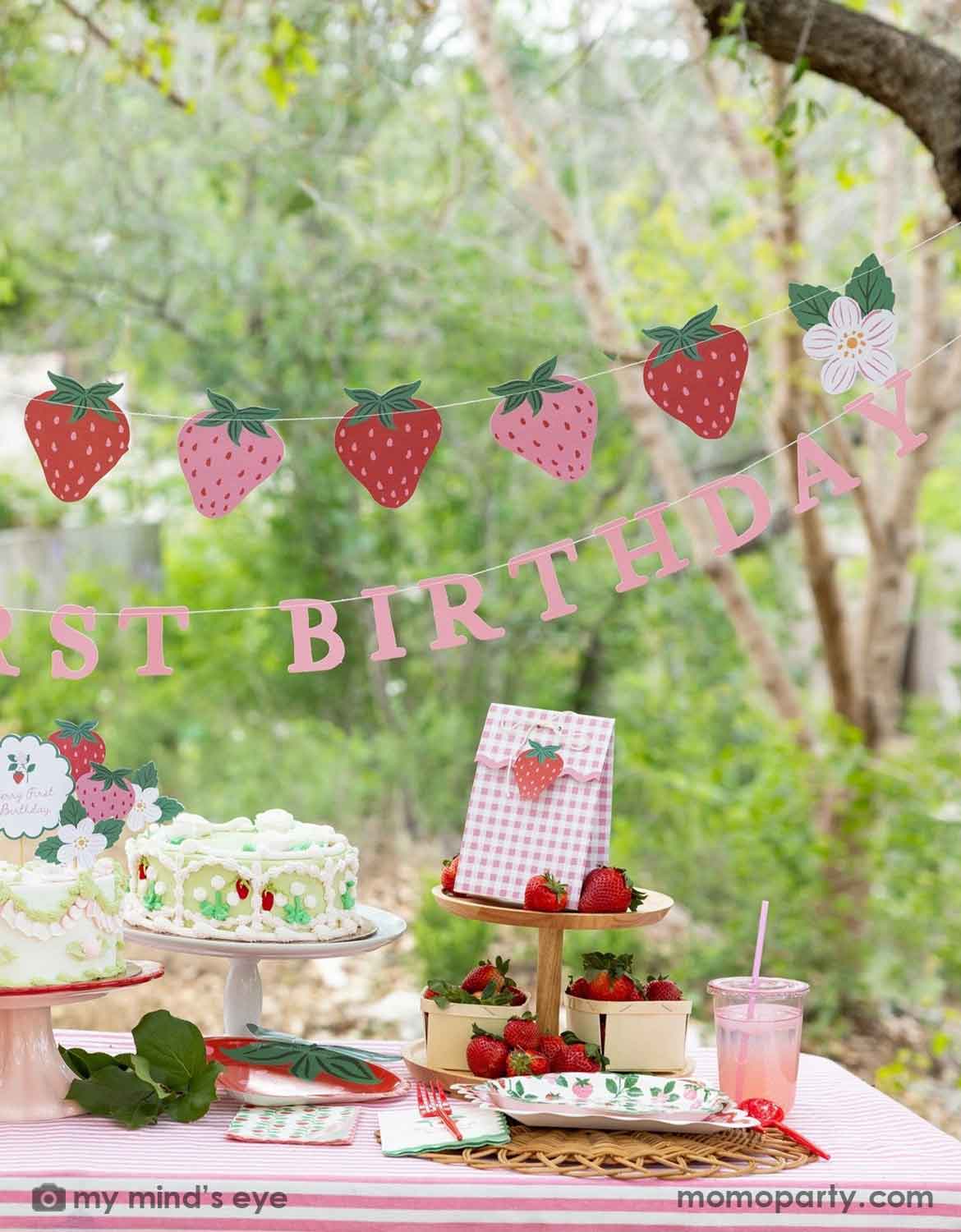 A Berry First strawberry-themed first birthday party setup outdoors, featuring various strawberry themed party supplies from Momo Party including a pink and red strawberry banner garland, a 'BERRY FIRST BIRTHDAY' banner, strawberry cake topper decorations on a vintage strawberry butter cake, and gingham party favor bags with strawberry cutouts, along with strawberry themed tablewares like plates, napkins and cups on a table with a blurred green garden background.