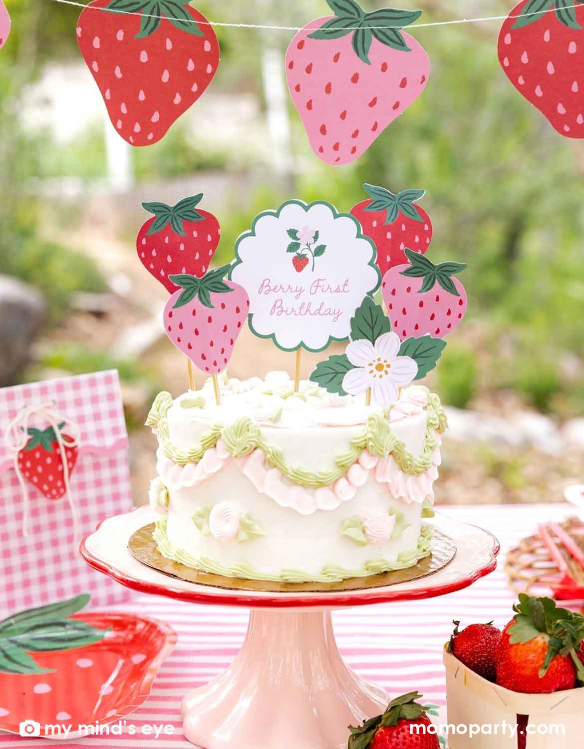 A strawberry-themed birthday cake on a pink cake stand stand, decorated with white frosting, green piped borders, and strawberry toppers read "Berry First Birthday" from Momo Party, set in an outdoor garden environment with a matching strawberry party garland hung above and tablewares of strawberry party plates, napkins and favor bags. 