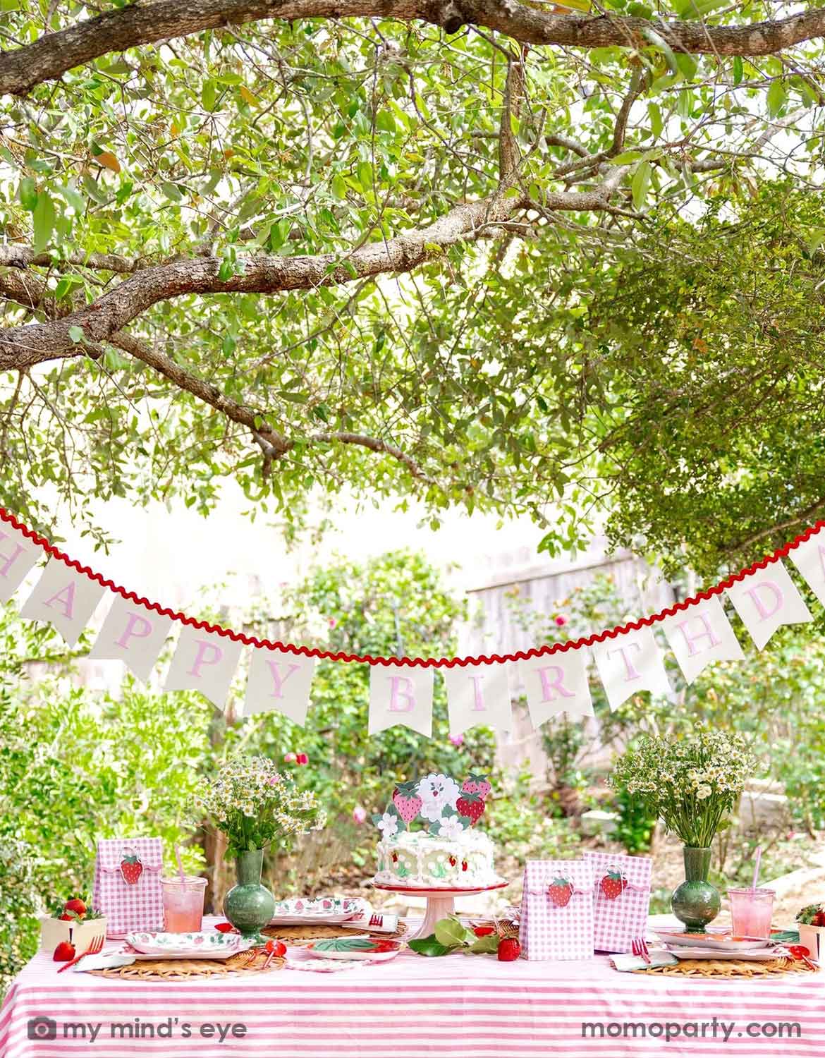An outdoor "Berry First" strawberry themed first birthday party setup under a leafy tree, with a pink-and-white striped tablecloth, strawberry-themed party supplies including plates, napkins, cups, party favor bags from Momo Party, along with a buttercream cake topped with strawberry toppers and a pink party banner reading 'HAPPY BIRTHDAY' hung above.