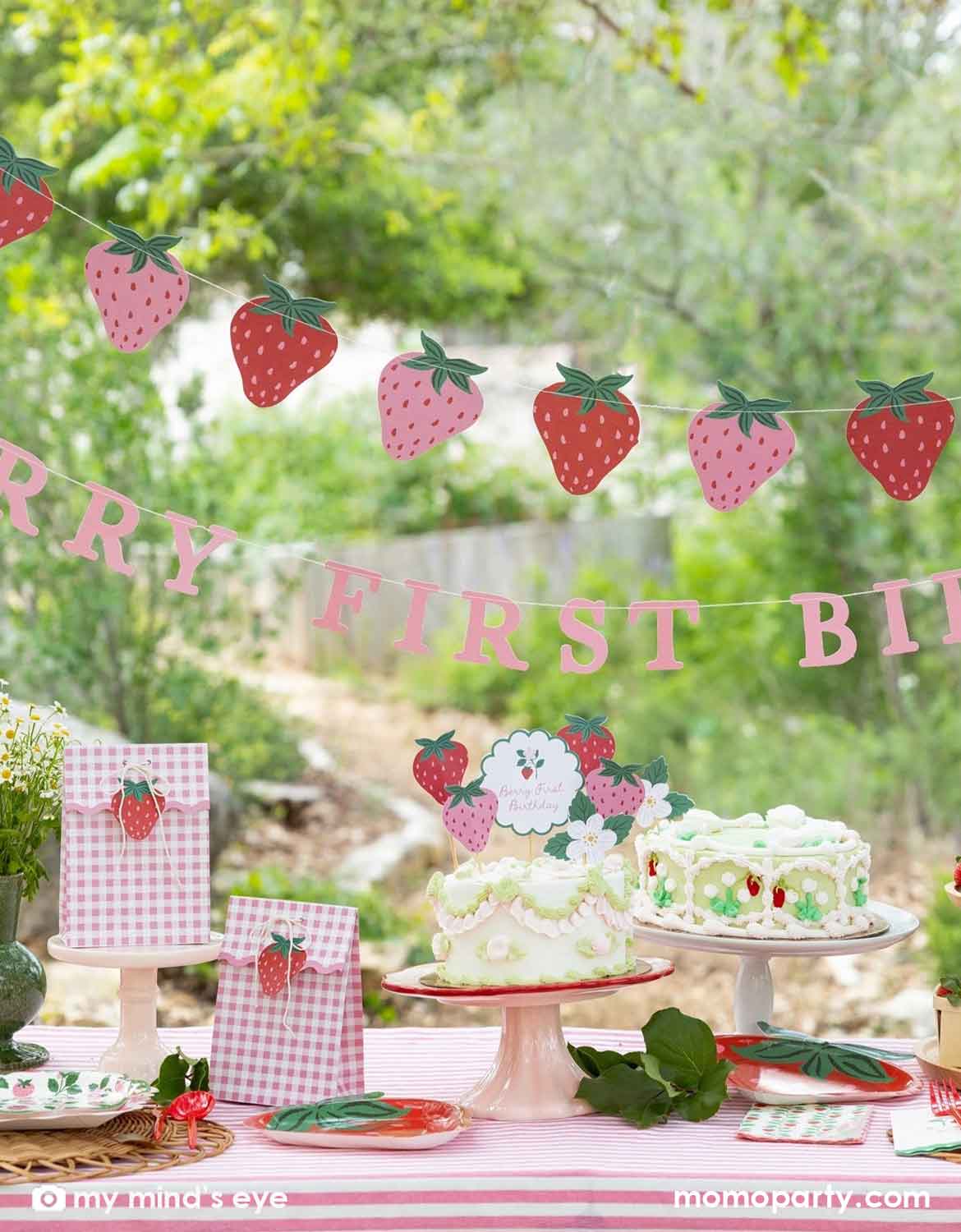 A Berry First strawberry-themed first birthday party setup outdoors, featuring various strawberry themed party supplies from Momo Party including a pink and red strawberry banner garland, a 'BERRY FIRST BIRTHDAY' banner, strawberry cake topper decorations on a vintage strawberry butter cake, and gingham party favor bags with strawberry cutouts, along with strawberry themed tablewares like plates, napkins and cups on a table with a blurred green garden background.