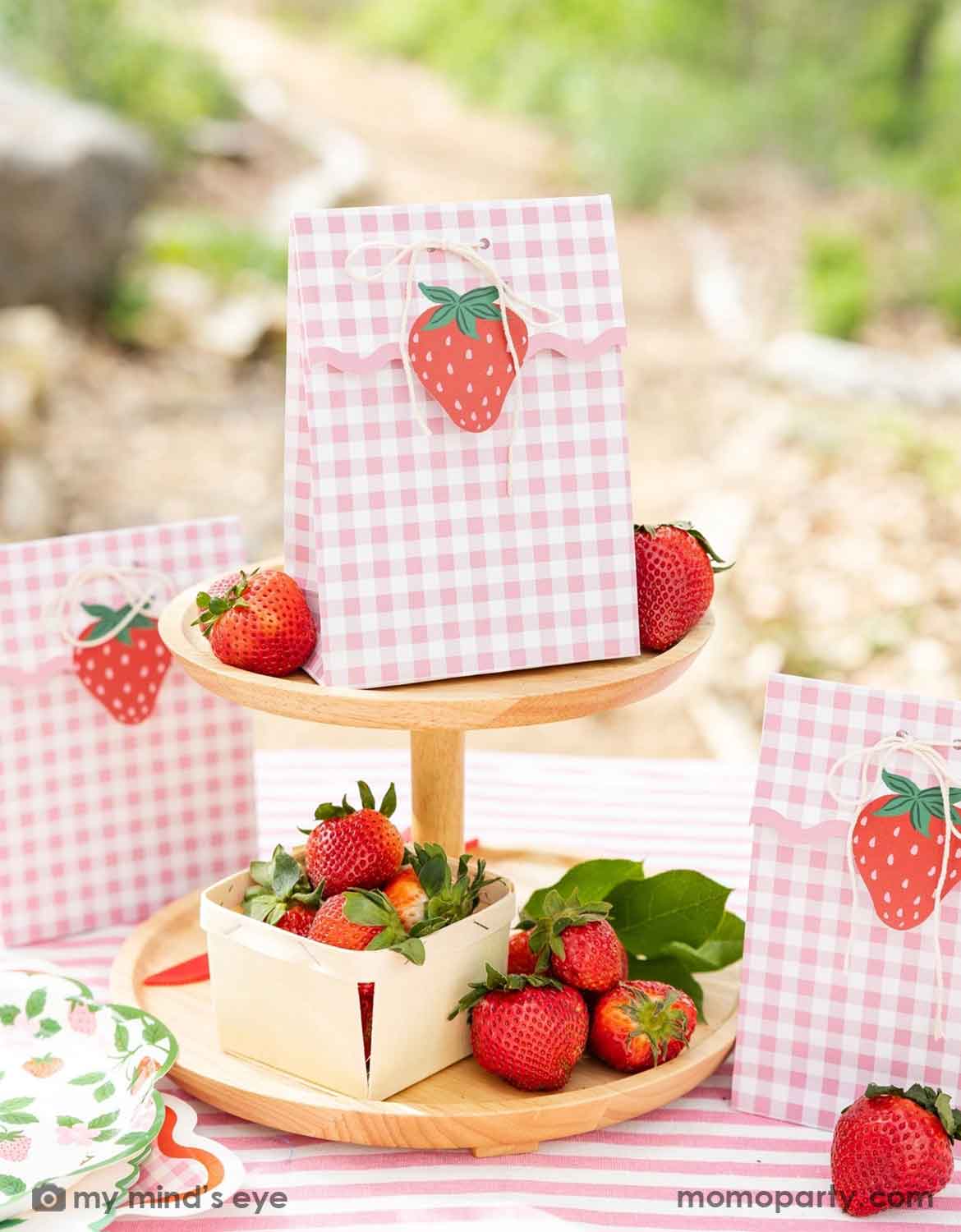 An adorable strawberry-themed party table with strawberry themed decorations and tableware from Momo Party, including pink gingham favor bags with strawberry cutouts, 9" strawberry and vine paper plates and dinner napkins stacked, and strawberries arranged on a wooden cake stand in an outdoor garden setting - a perfect idea for girls "Berry First"  strawberry themed first birthday celebration or any strawberry themed parties.