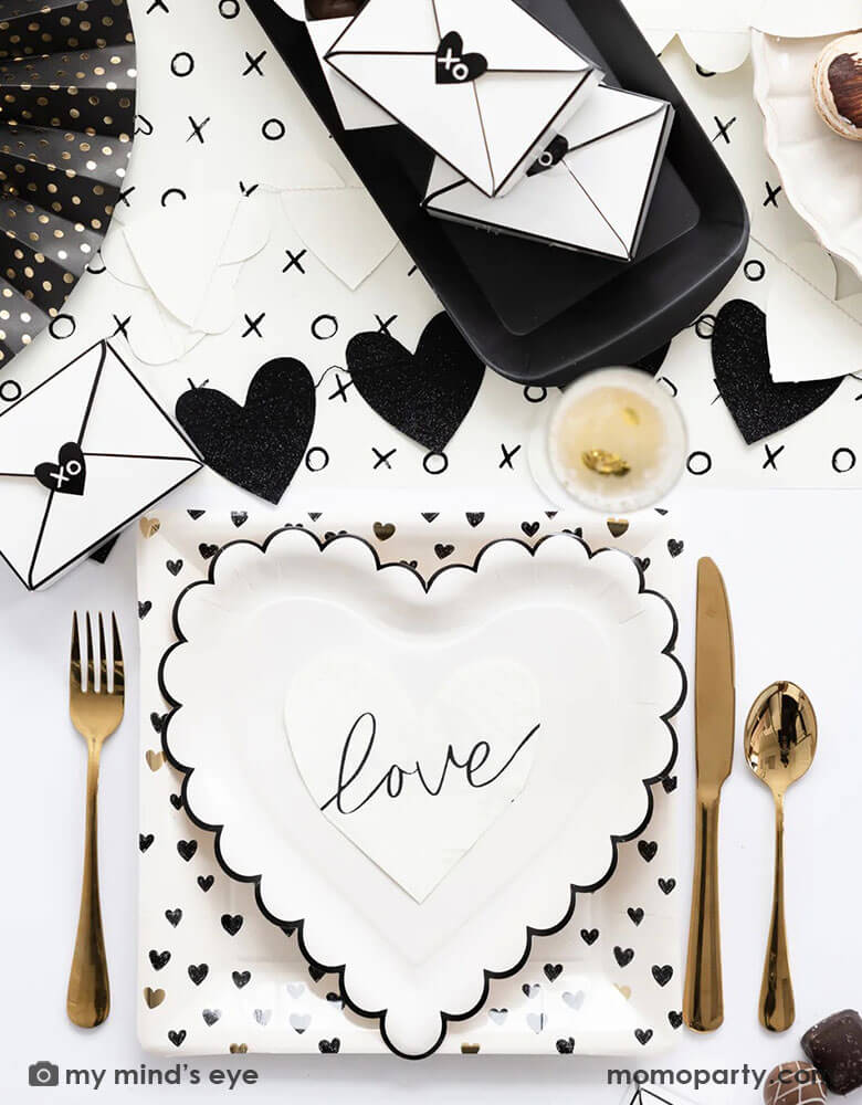 A black and white sophisticated Valentine's Day party tablescape featuring Momo Party's Valentine's Day party supplies including the XOXO paper table runner, heart shaped plates and black heart scattered dinner plates.