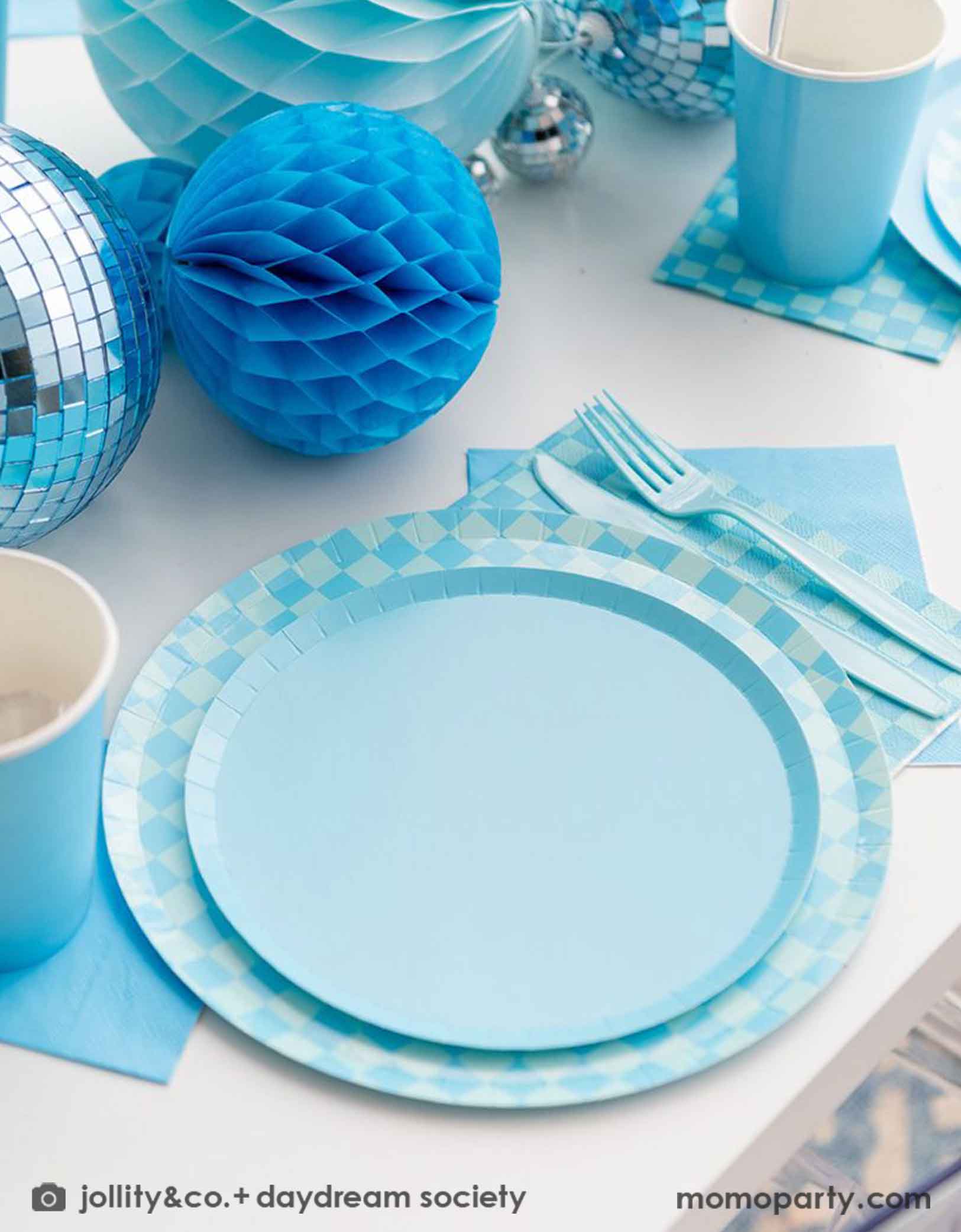 A blue themed party table features Momo Party's Blue and Check it! Blue Checkered Tableware Collection by Jollity & Co. including 10" blue checkered dinner plates, 8" blue dessert plates, 6.5" blue checkered large napkins, 5" blue small napkins, blue plastic utensils and solid blue party cups, with blue honeycomb and disco ball decorations as the centerpiece for the party table.
