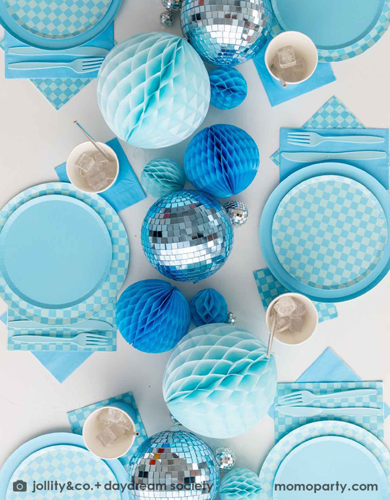A overhead shot of a blue themed party table features Momo Party's Blue and Check it! Blue Checkered Tableware Collection by Jollity & Co. including 10" blue dinner plates, 8" blue checkered dessert plates, 6.5" large napkins, blue plastic utensils and solid blue party cups, with blue honeycomb and disco ball decorations as the centerpiece for the party table.