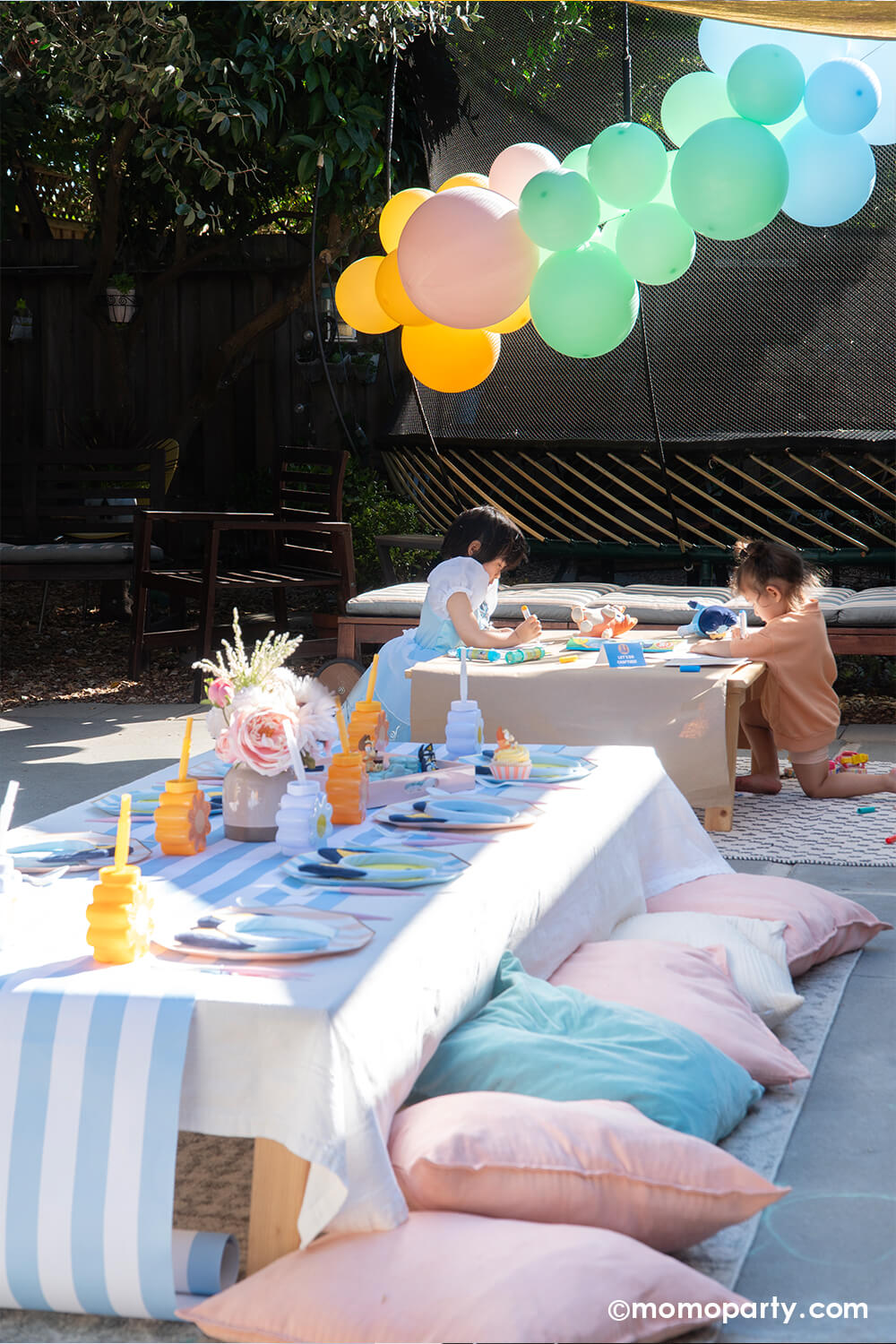 A kid's Bluey themed birthday party outdoor setting styled by Momo Party with a kid's picnic table, soft colored decorative pillows. On the table it was set with Bluey inspired party tablewares from Momo Party in stripe and daisy flower designs. Behind the picnic table two preschool aged girls doing arts and crafts table under a pastel colored balloon arch hung on the trampoline.