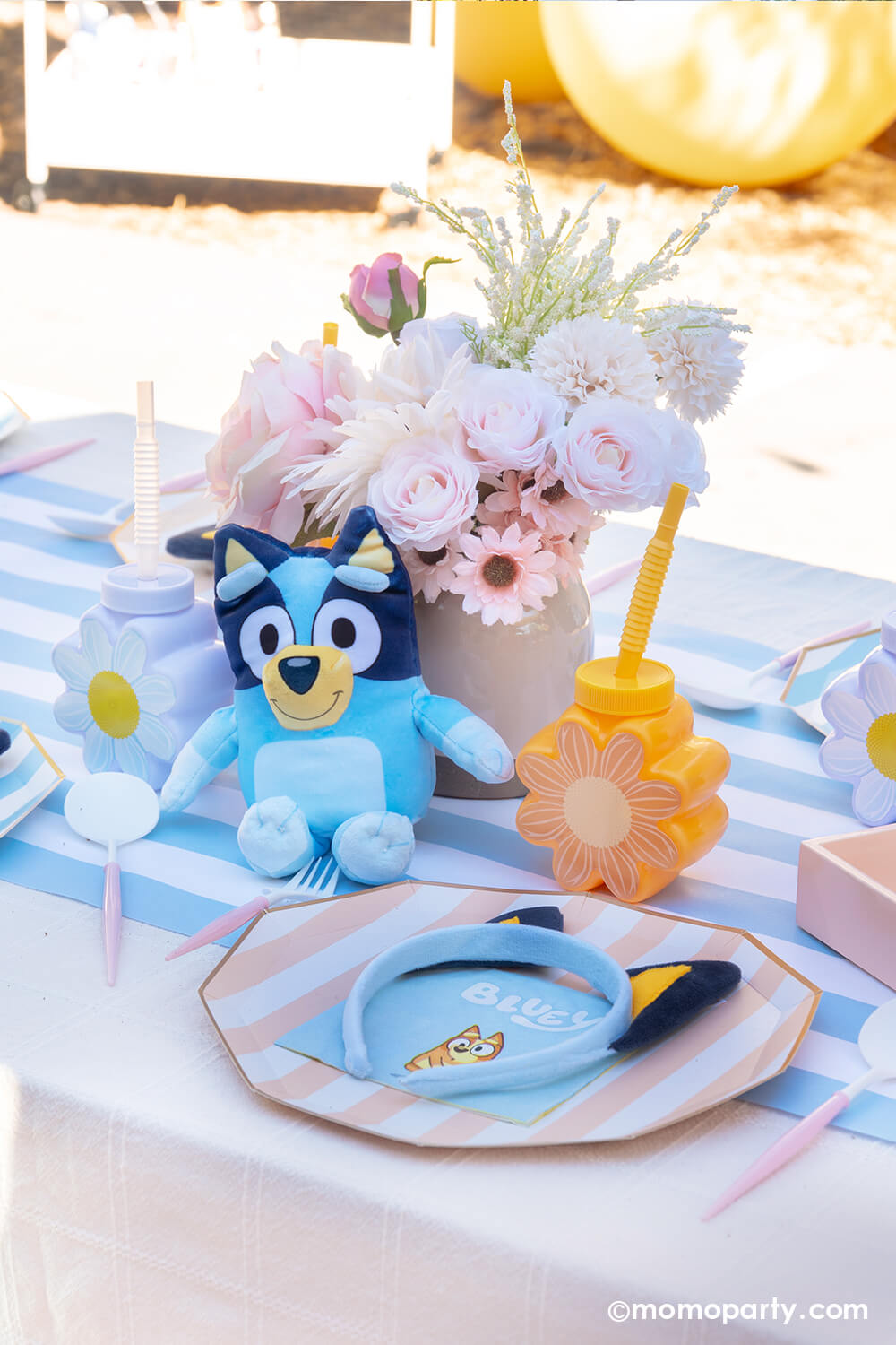 An adorable kid's picnic party table setting with Bluey themed various tablewares from Momo Party including peach and summer blue striped dinner plates, light blue striped table runner, daisy shaped tumbler cups in peach and white, Bluey small napkins, Bluey decorative headbands. Along with a small Bluey plush toy, floral bouquet decorations, it's a perfect table setting idea for a modern Bluey themed birthday celebration.