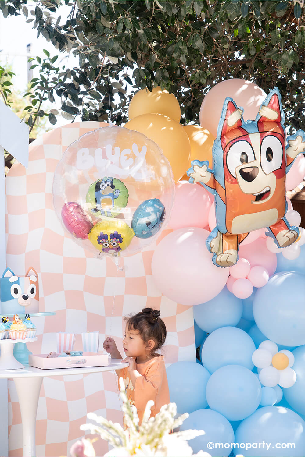 A pre-school aged girl standing in front of a wavy peach checkered party backdrop board with Bluey themed balloon garland in pastel peach, pink and blue adorned with daisy balloons. The balloon garland is adorned with the 32" x 23" Bluey bingo shaped foil balloon and Bluey insiders balloons with Bluey characters in different colors. With a Bluey and Bingo shaped birthday cake and desserts on the table, this adorable setting is perfect for your Bluey-loving child's birthday celebration.