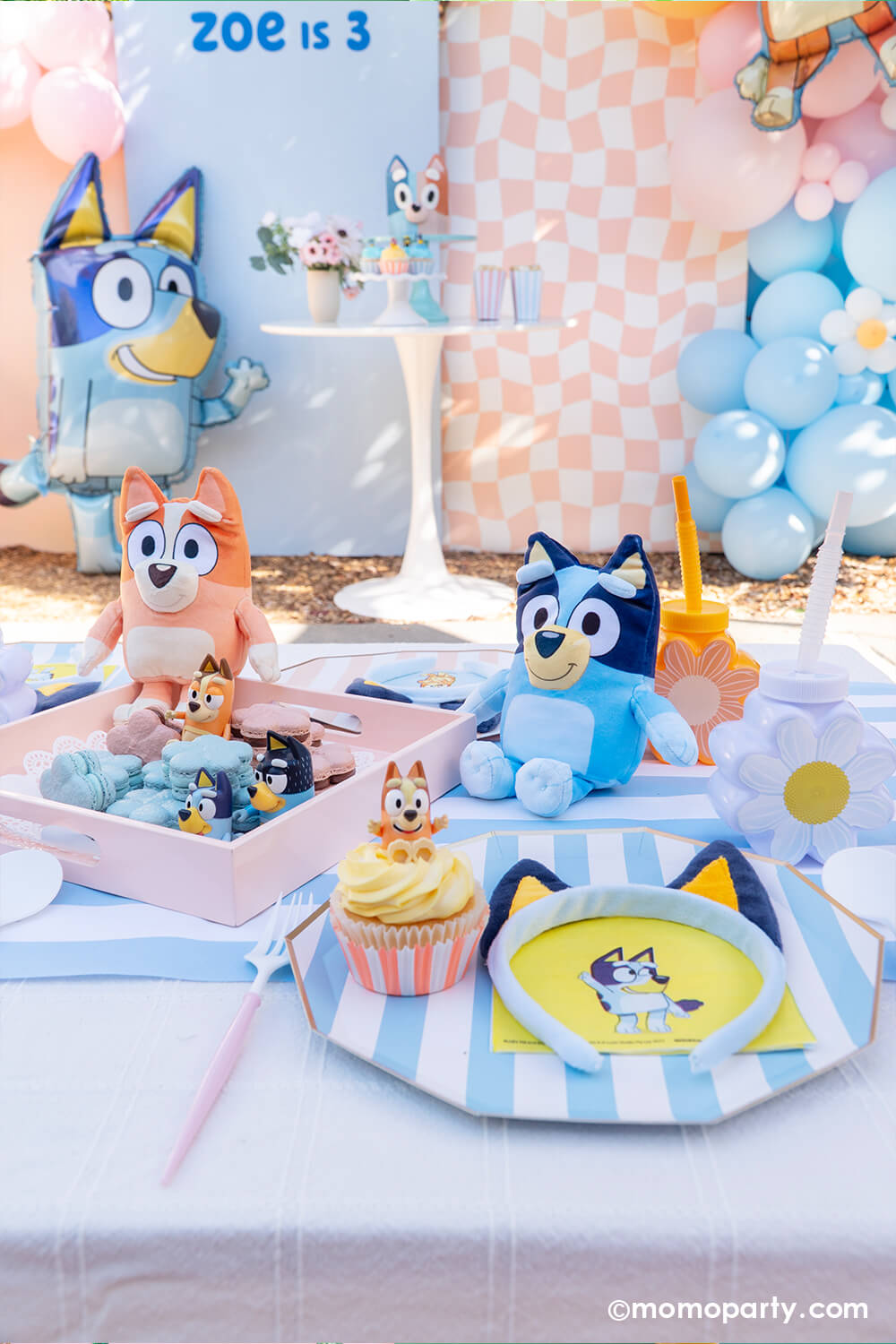 A kid's Bluey themed birthday party set up by Momo Party featuring pastel blue and peach backdrop boards adorned with a soft colored balloon garland. In the front, there's the jumbo 36" Bluey AirWalkder foil balloon tanding next to a dessert table with a Bluey themed sweets. In the front set a kids picnic table with Bluey themed tableware including blue striped plates, Bluey napkins, daisy shaped party cups and Bluey headbands from Momo Party. Along with Bluey & Bingo stuffies, cupcakes and daisy macarons.