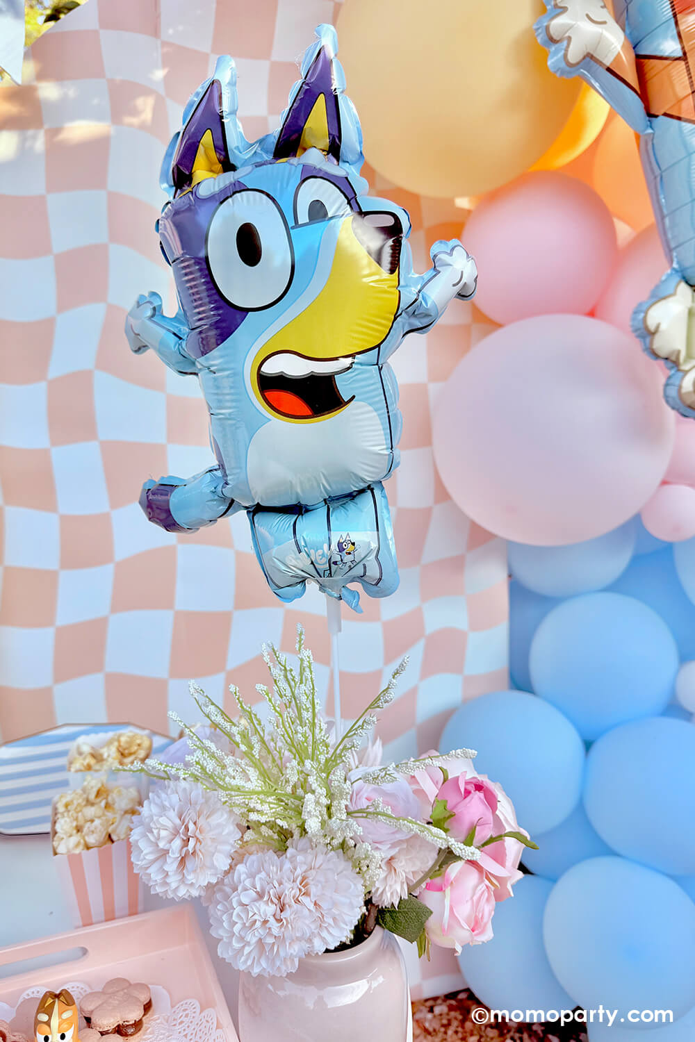 Momo Party's 10" x 13" mini Bluey shaped foil balloon with a colorful balloon garland of pastel pink, blue and peach, along with flower bouquet on a peach checkered background.