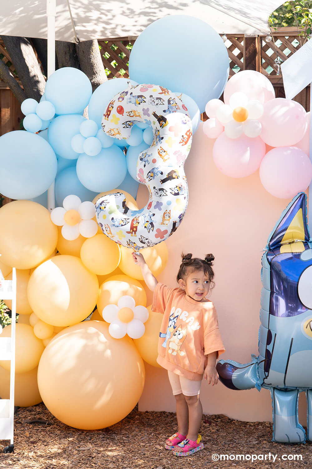 A little gril in Bluey clothes standing in front of a pastel colorful balloon arch in peach, baby blue and pink adorned with daisy shaped balloons with a number '3' balloon with Bluey cartoon characters, next to a jumbo Bluey shaped AirWalker foil balloon from Momo Party.